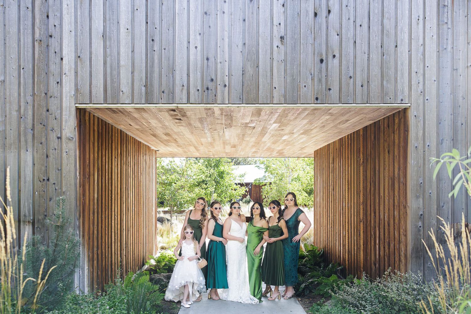Bride and bridesmaids in green dresses posing under a rustic wooden archway surrounded by lush greenery.