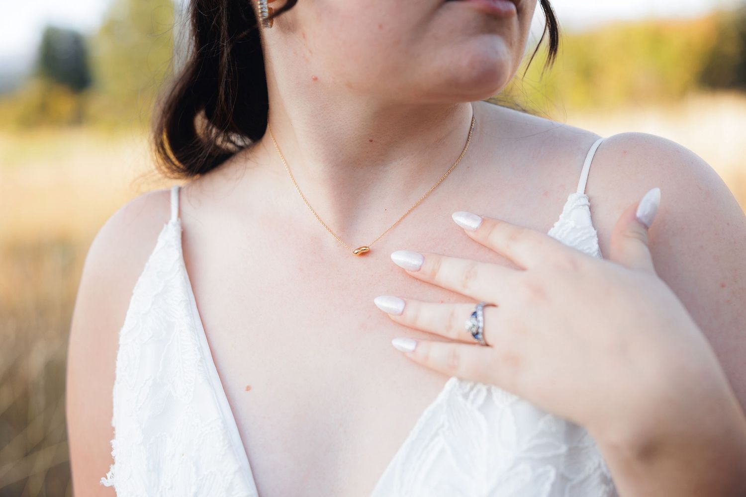Bride in white dress showcasing elegant engagement ring and delicate necklace outdoors.