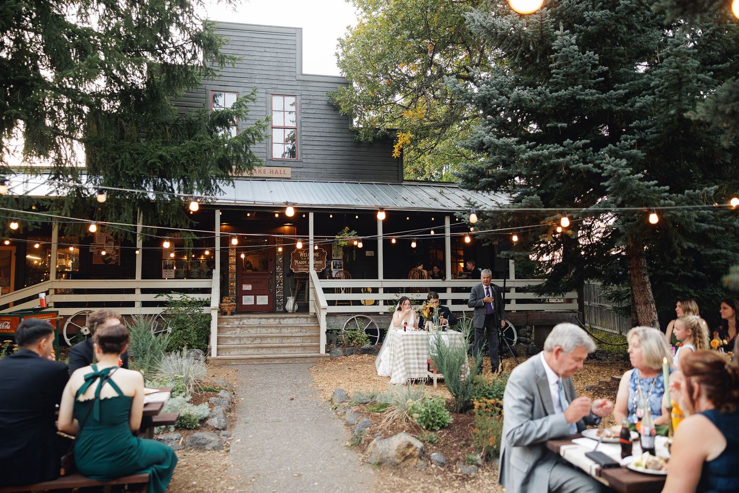 Outdoor wedding reception at rustic barn venue with string lights, guests dining al fresco at wooden tables surrounded by lush trees.