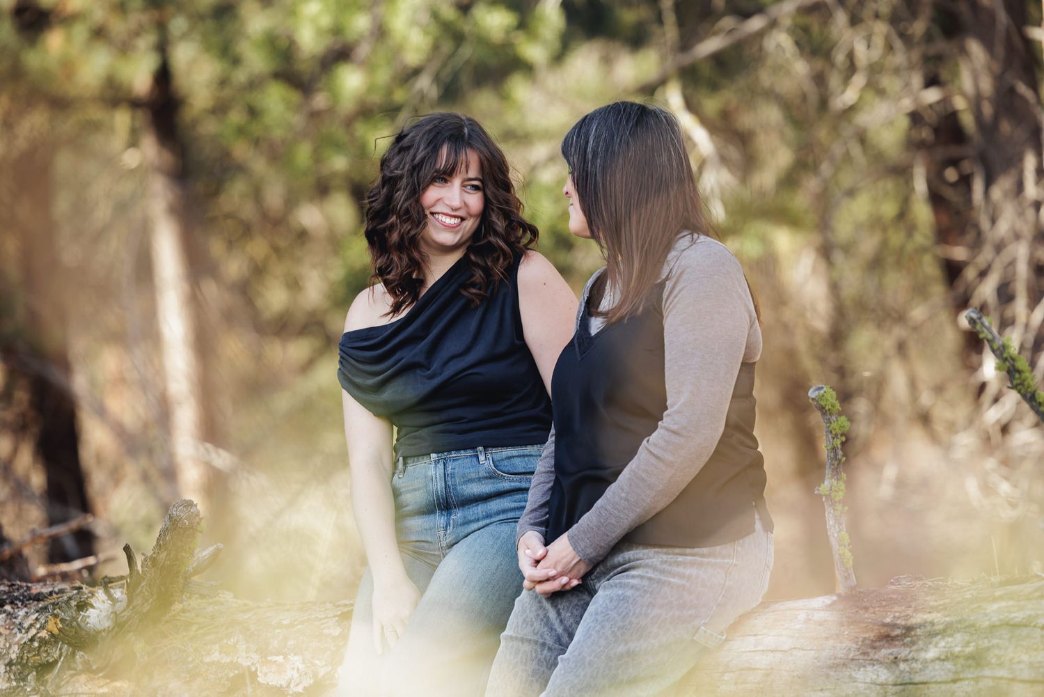 Two women sitting together laughing outdoors in a sunlit forest setting, wearing casual tops and jeans.