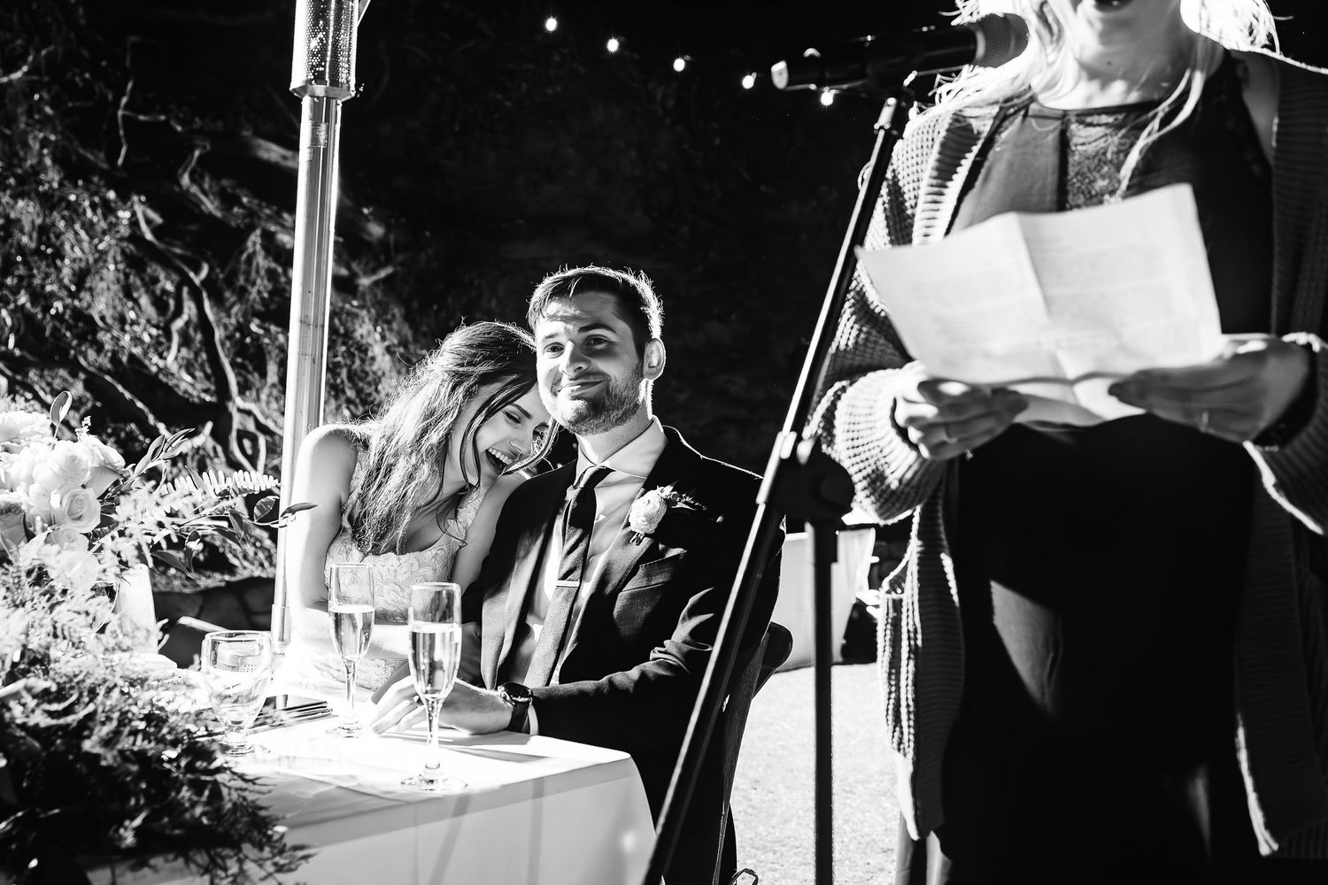 A black and white photo of a couple at a wedding ceremony while an officiant reads from a paper at a podium.