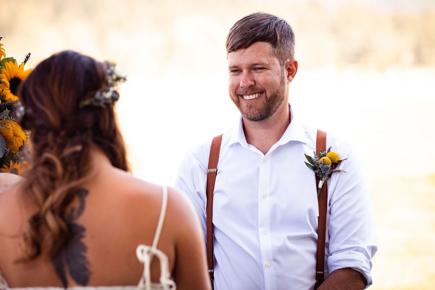 Happy groom in a white shirt and suspenders smiles during outdoor wedding ceremony with sunflowers and warm lighting.