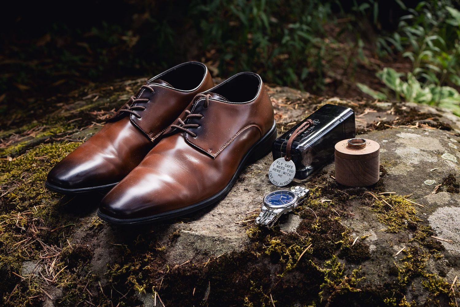 Brown leather dress shoes with watch and accessories arranged on mossy stone surface in natural setting.