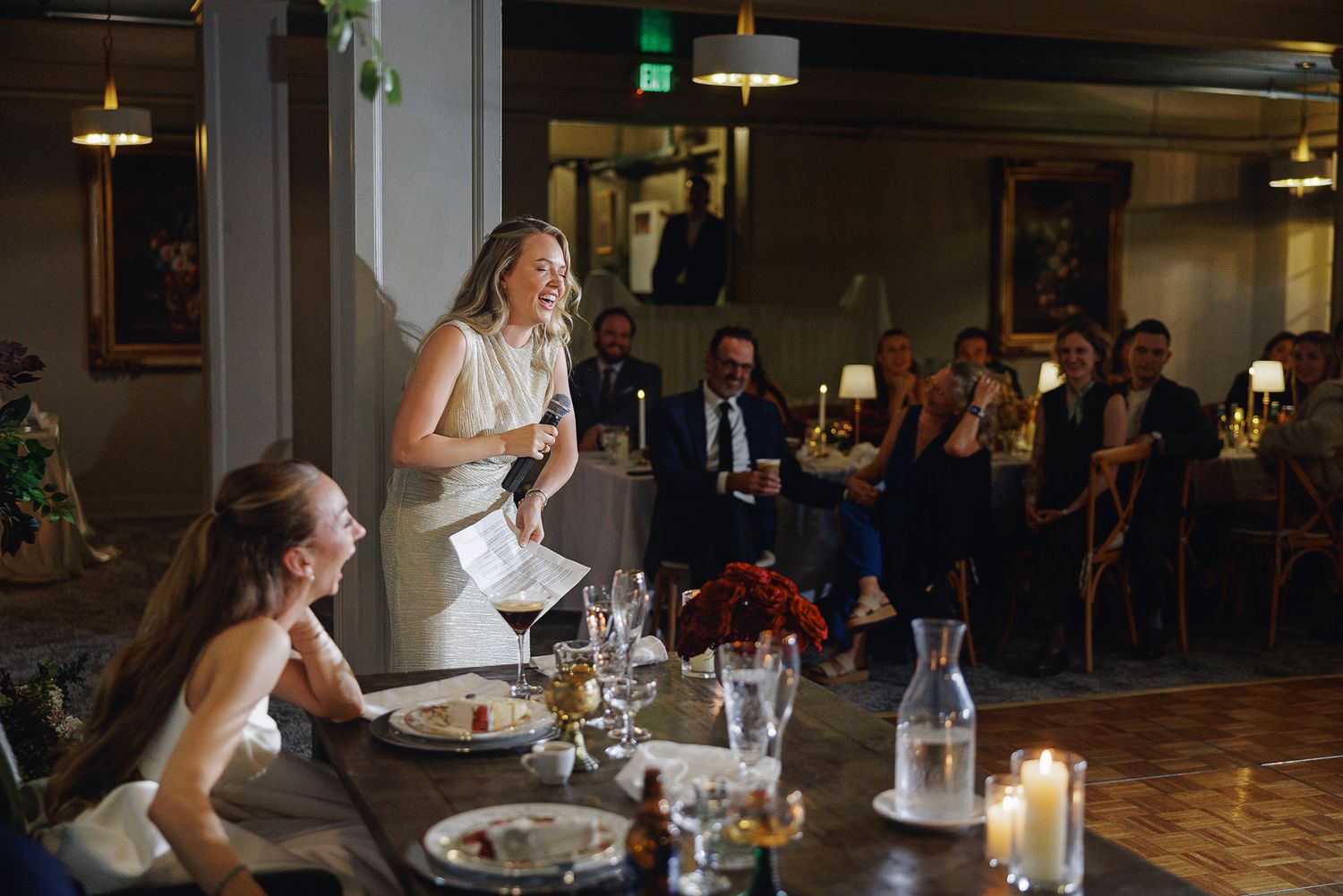 Blonde woman giving a speech at an elegant wedding reception dinner while guests applaud.