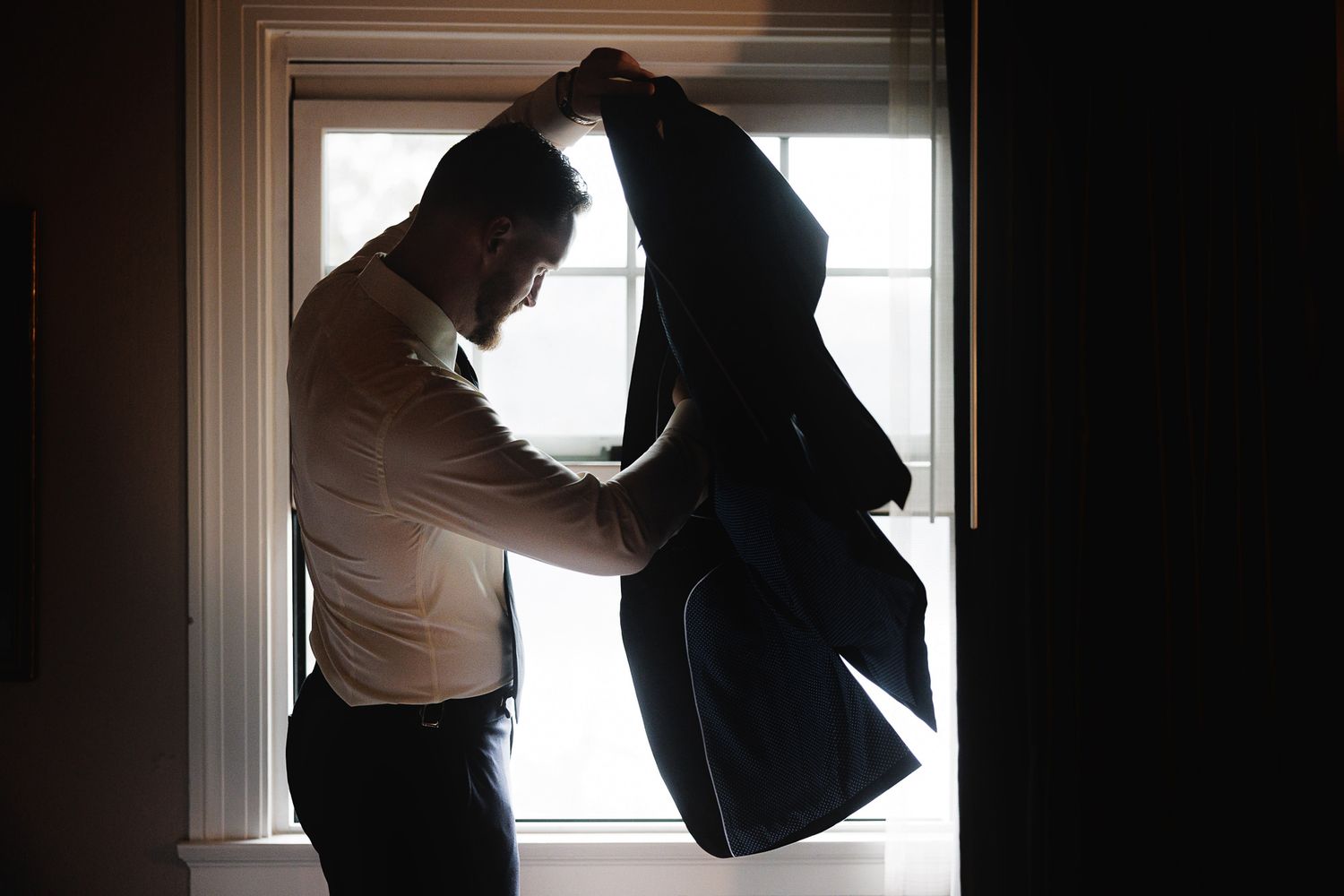 A groom putting on his suit jacket in silhouette by a bright window while getting ready for his wedding day.
