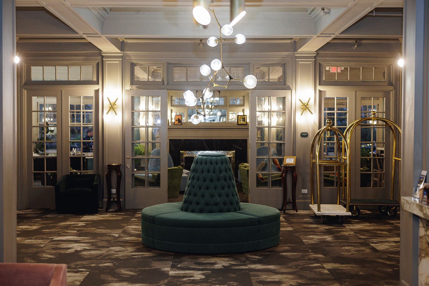 Elegant hotel lobby with tufted circular green sofa, chandelier, fireplace, French doors, and gold luggage cart on patterned carpet.
