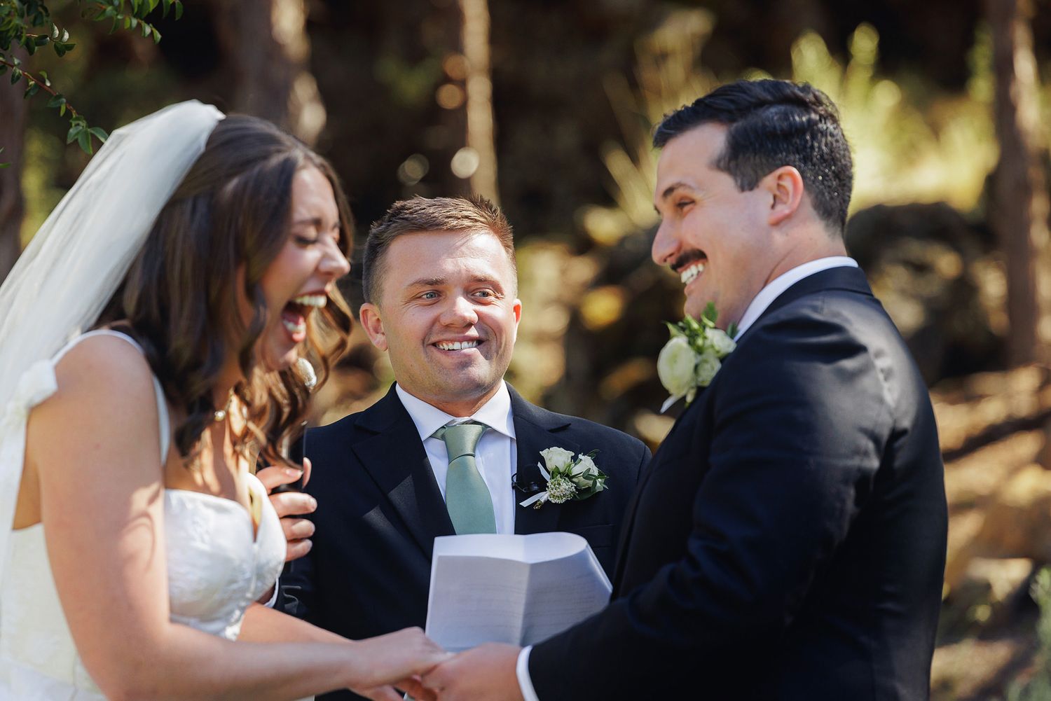 Bride and groom laughing with officiant during outdoor wedding ceremony in forest setting.