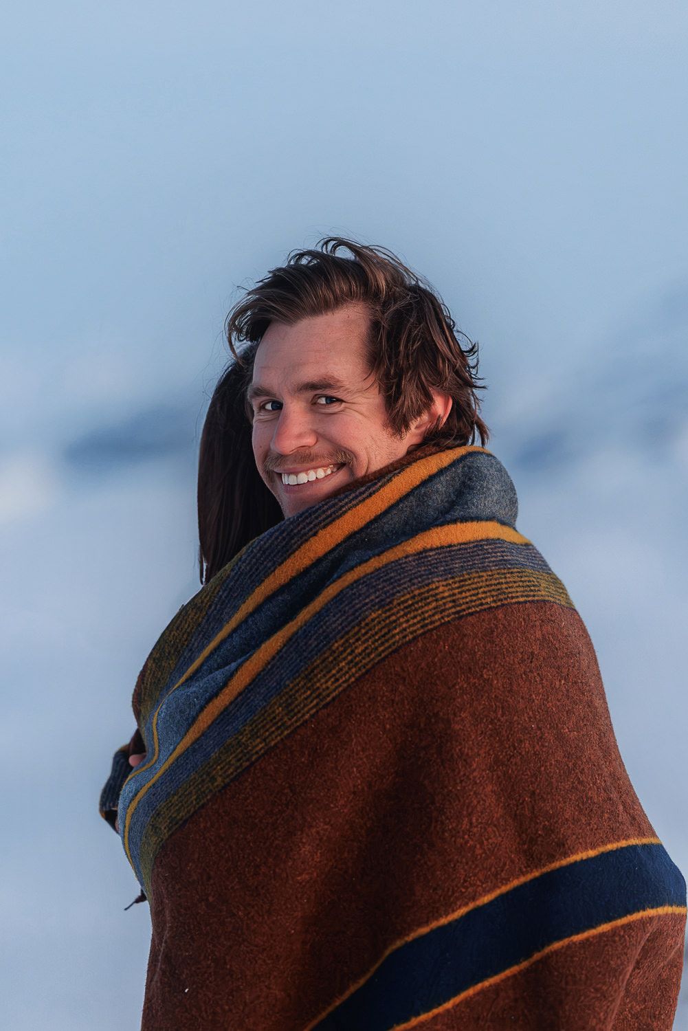 Someone wrapped in a brown blanket with orange stripes smiles against a blue winter sky background.