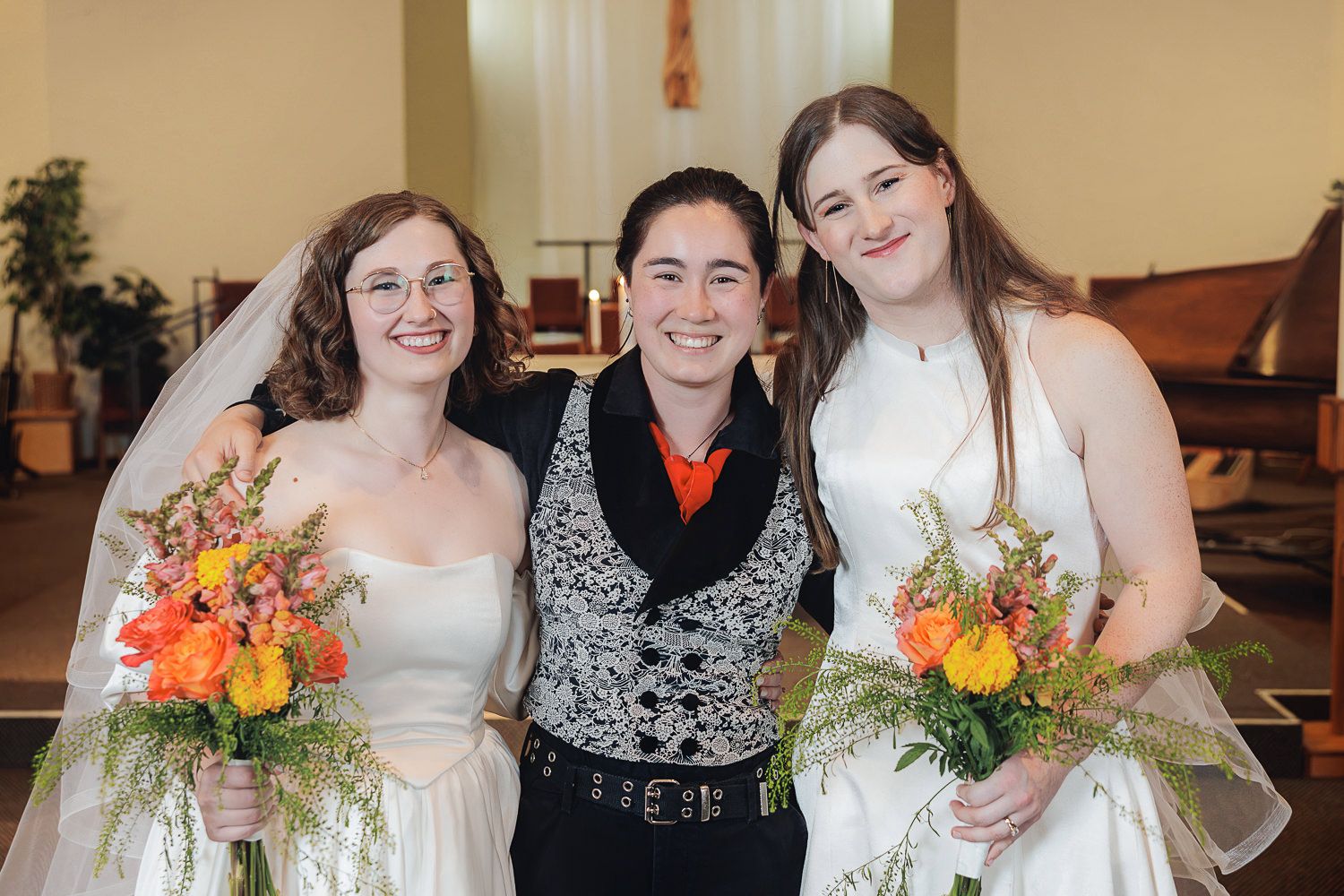 Three people pose together at a wedding celebration with orange and yellow floral bouquets.