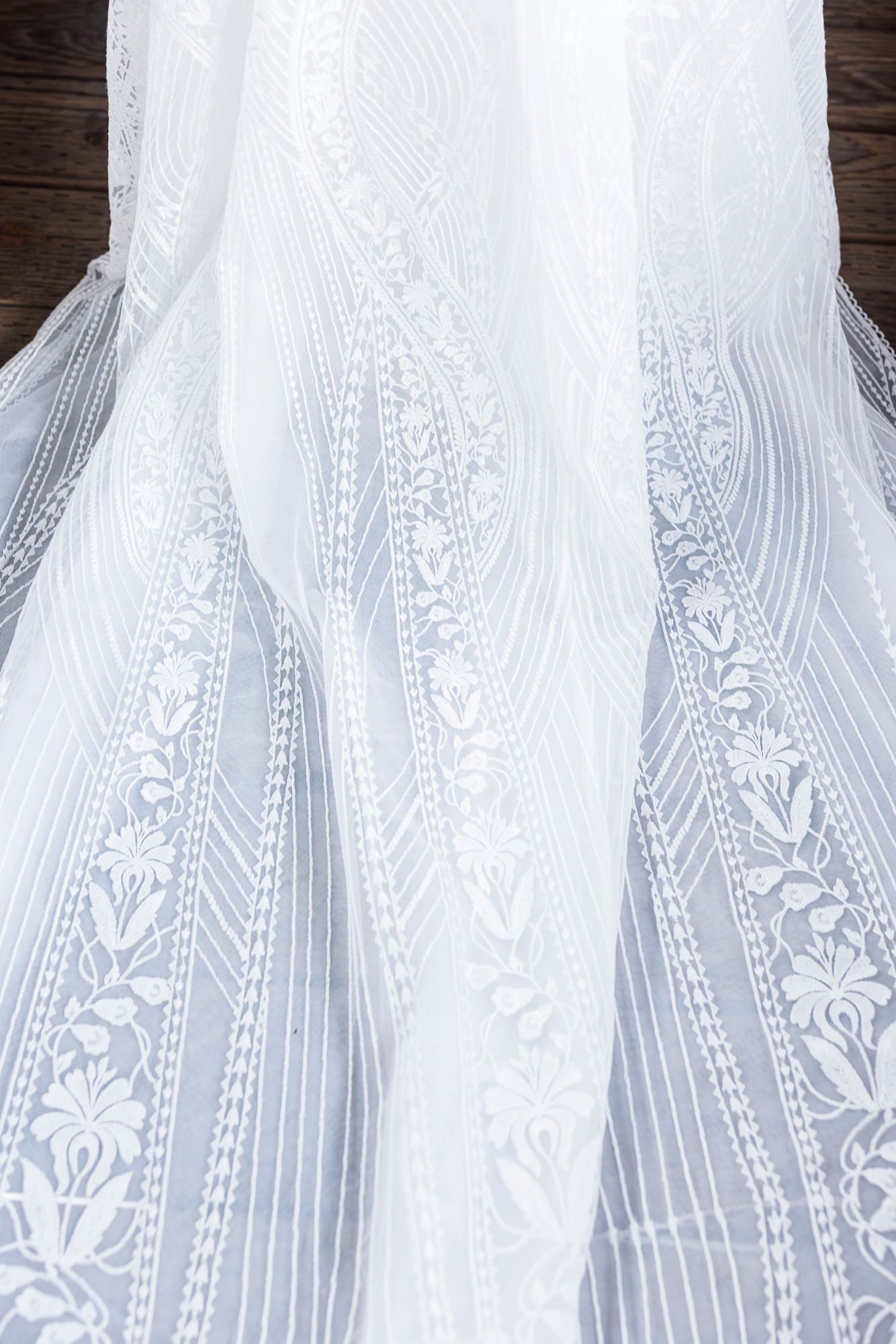 Close-up detail of delicate white lace and embroidered floral patterns on a wedding dress fabric against dark wood.