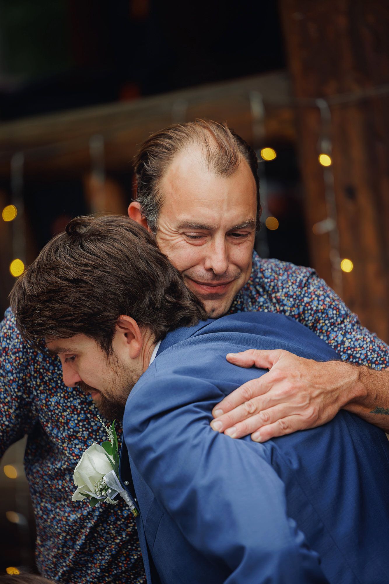 Two men share an emotional embrace at a wedding reception with twinkling lights in the background.