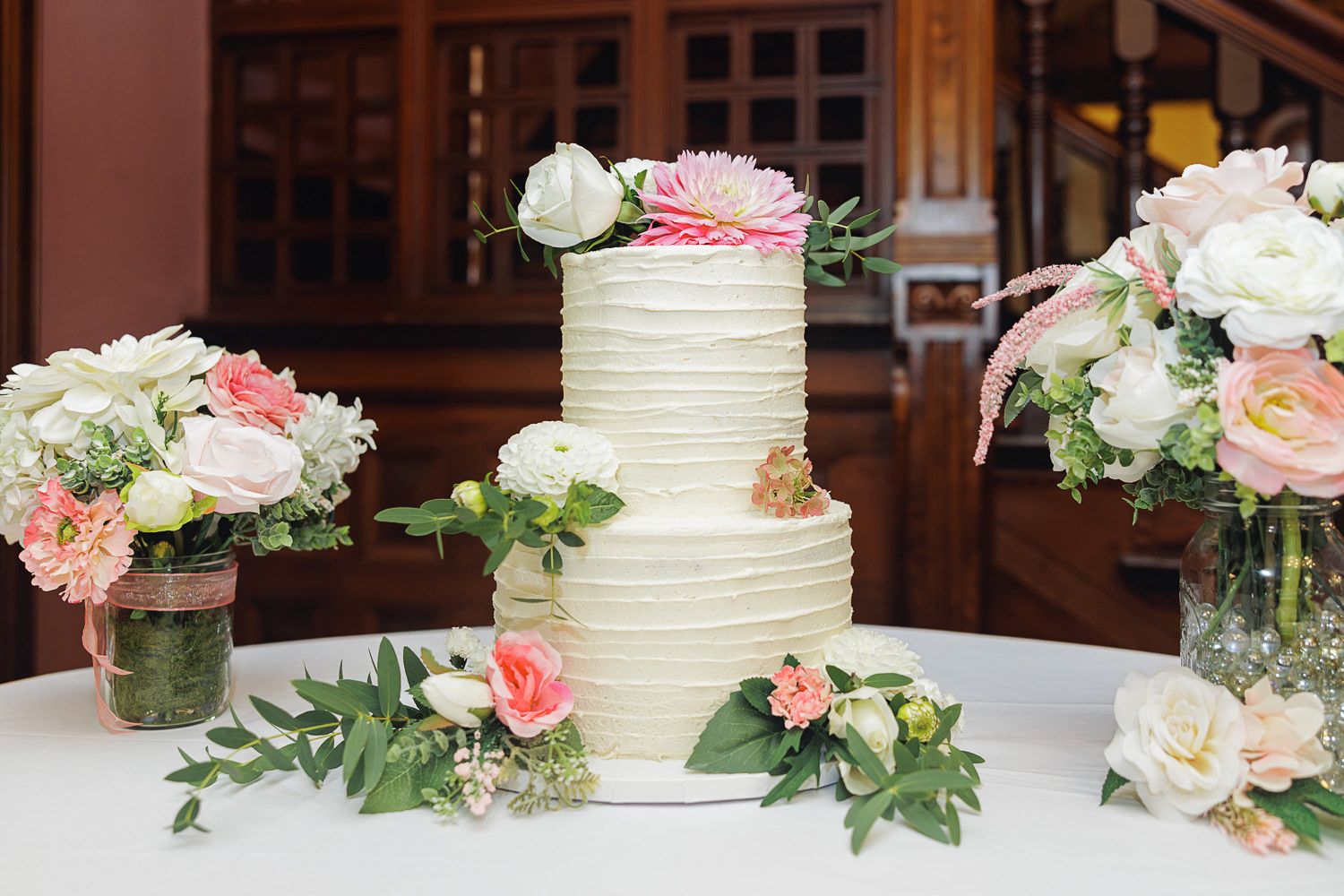 White wedding cake decorated with pink and peach roses and greenery sits on display at an elegant reception setting.