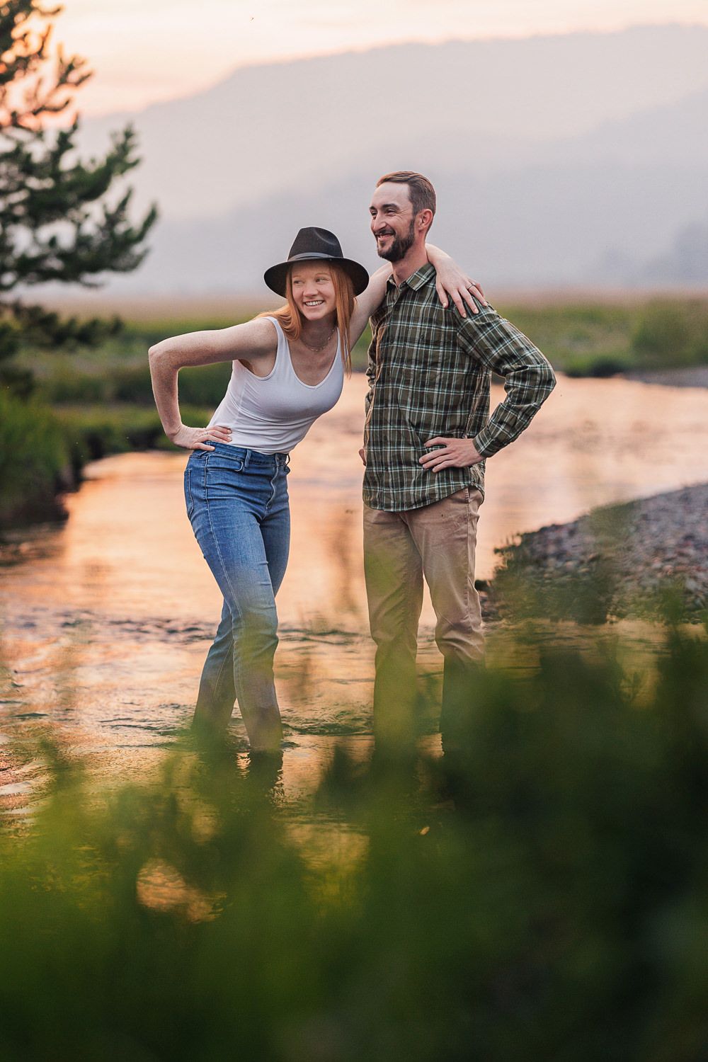 A couple laughs and dances together by a lake during golden hour sunset while wildflowers bloom in the foreground.