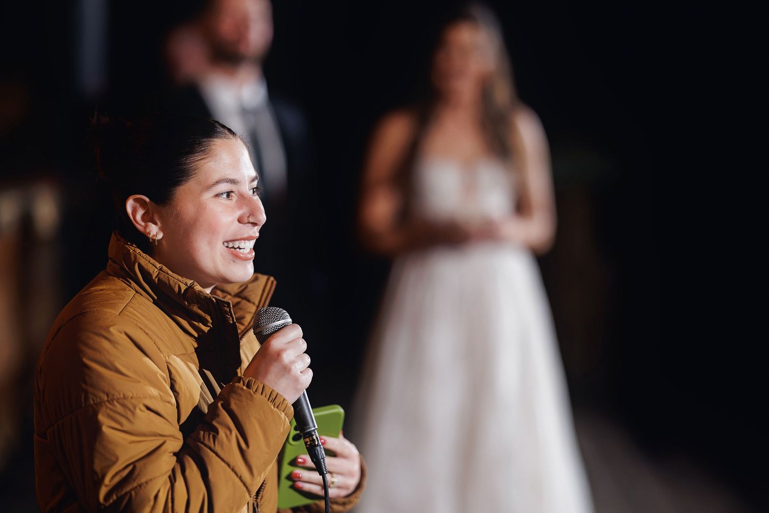 Woman in yellow jacket speaking into microphone at wedding reception with bride and groom blurred in background.
