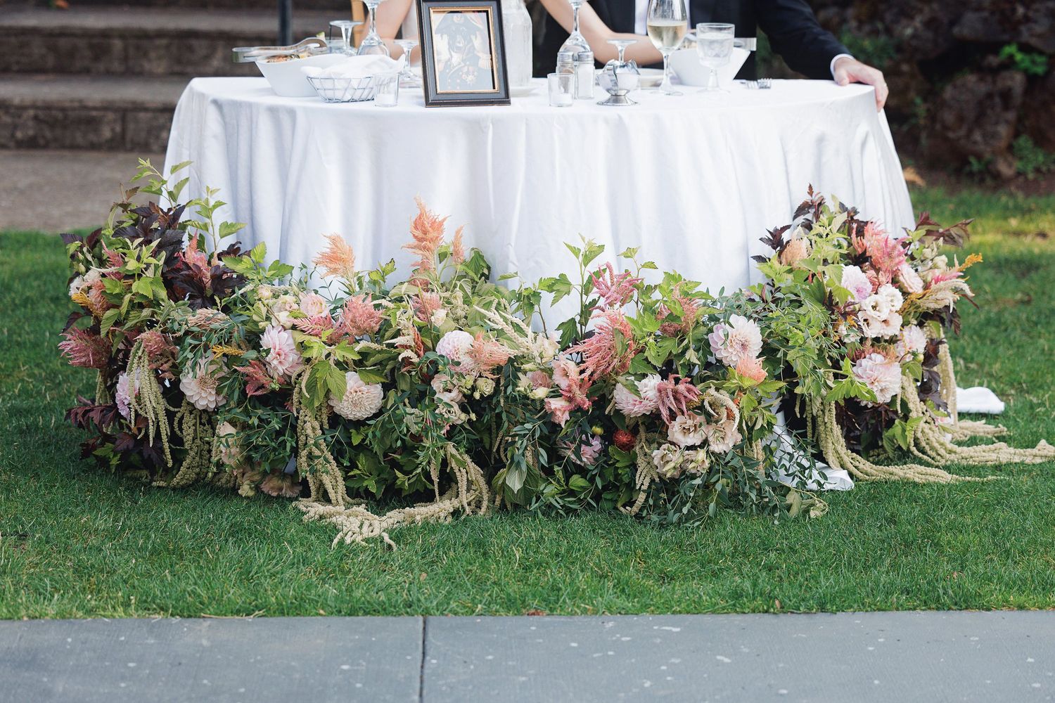 Elegant wedding sweetheart table with lush floral arrangements of pink and mauve blooms on green grass.