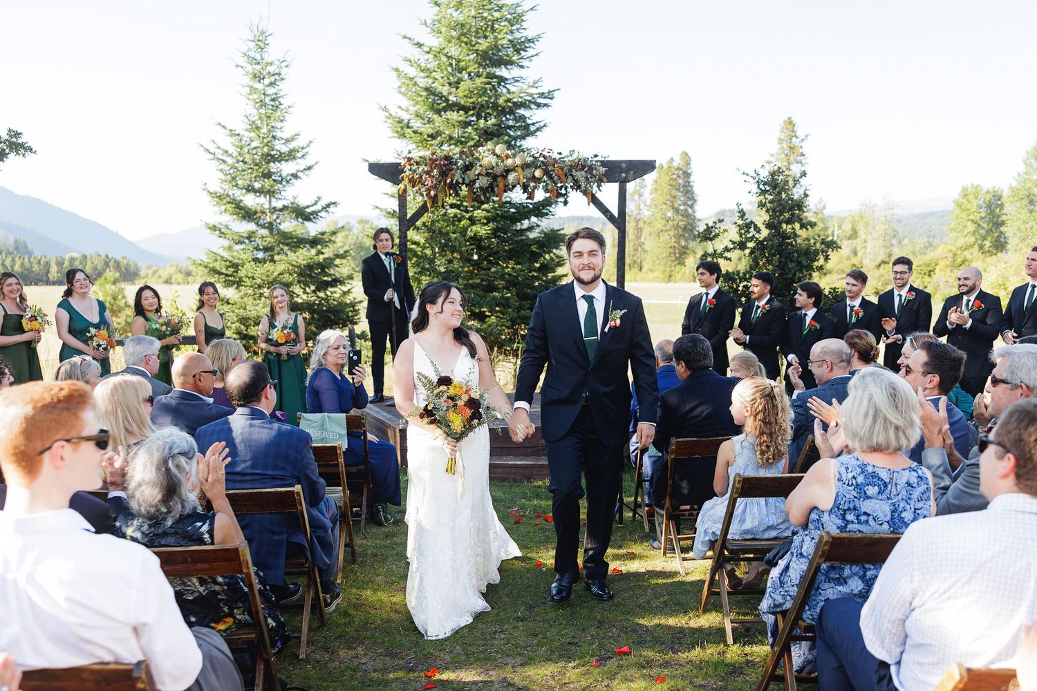 Bride and groom walk down outdoor aisle at mountain wedding ceremony with guests applauding on a sunny day.
