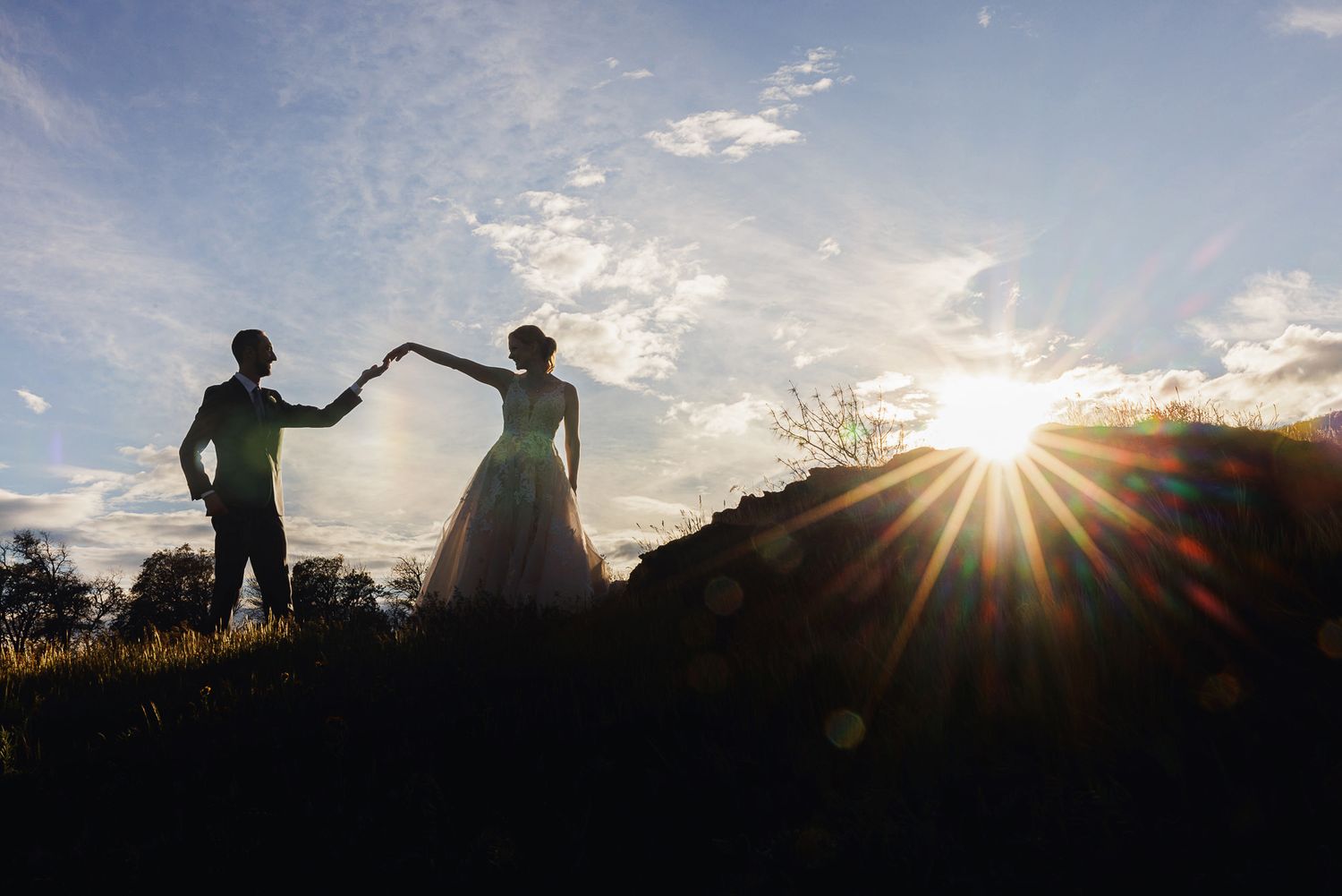 Silhouette of a bride and groom holding hands at sunset with dramatic lens flare and blue sky backdrop.