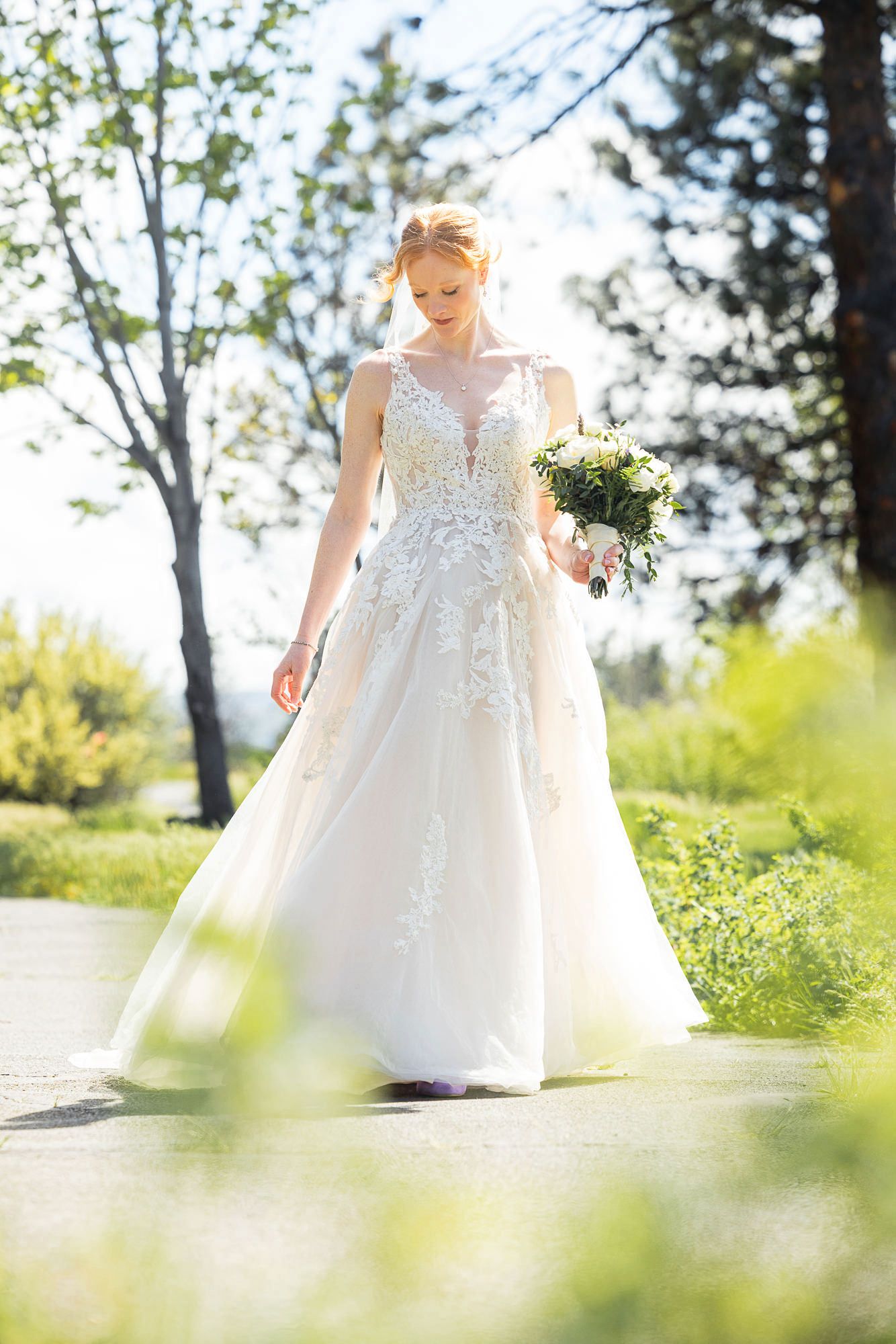Bride in white lace A-line gown holding bouquet walks outdoors on sunny path surrounded by green trees.