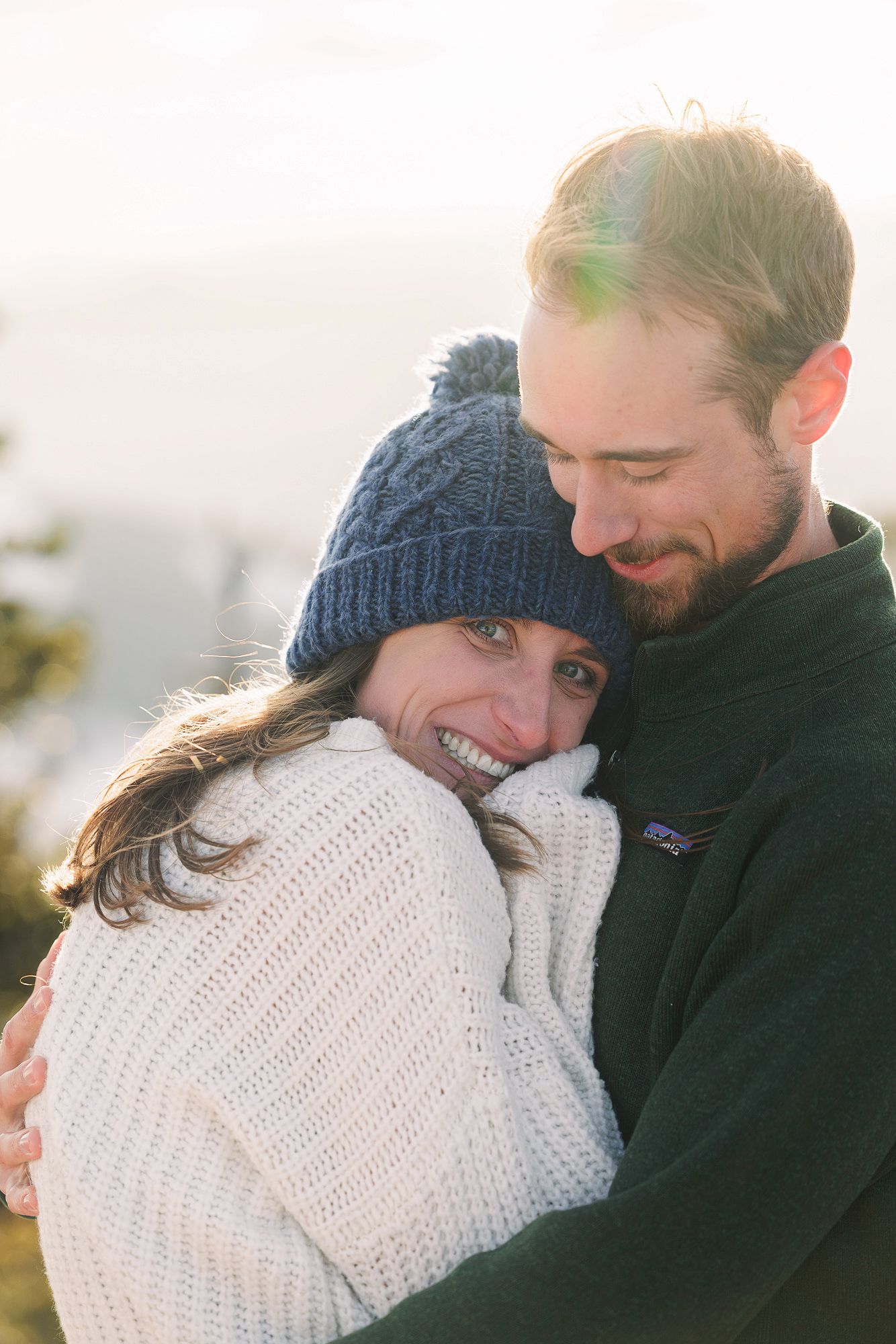 A smiling couple embraces outdoors in winter, woman wearing knit hat and white sweater, man in dark fleece jacket.