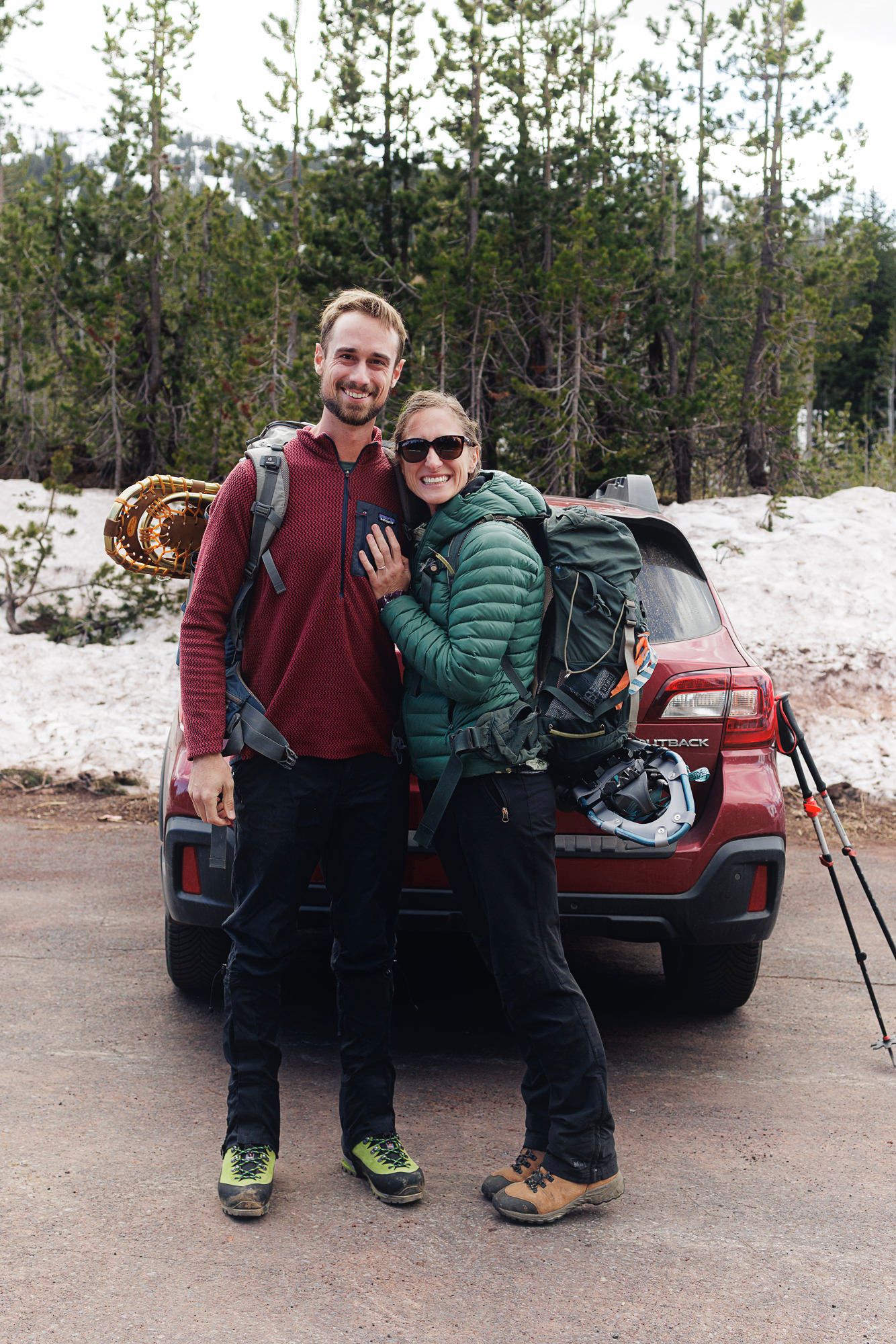 A couple in hiking gear smiles together in front of a red SUV surrounded by snowy forest terrain.