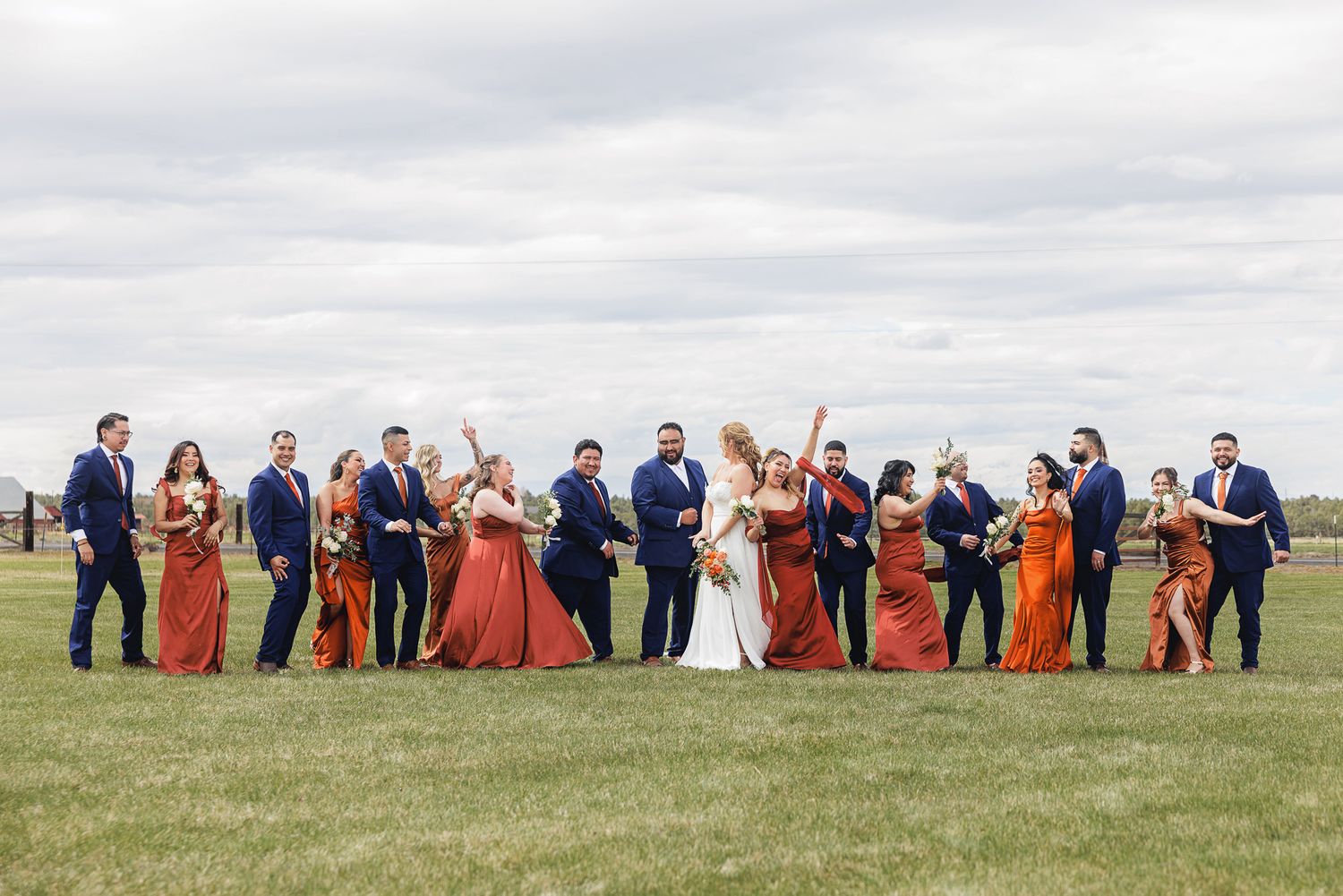Wedding party in navy suits and rust bridesmaid dresses celebrating outdoors on green grass under cloudy sky.