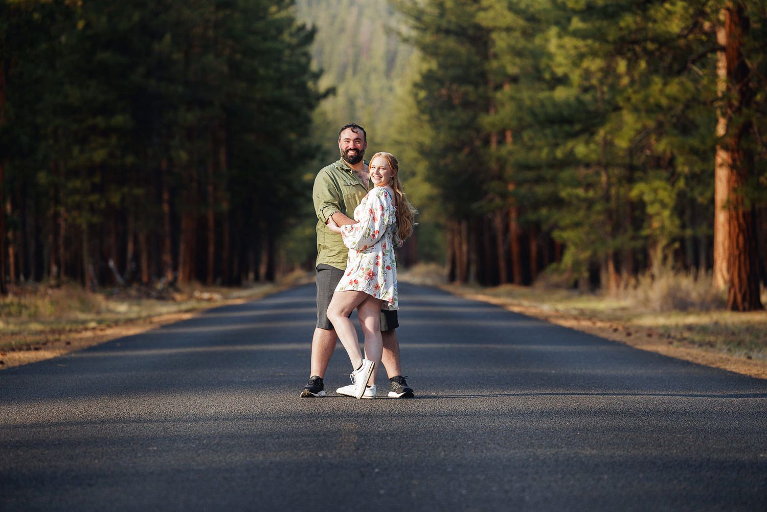 Couple dancing on a tree-lined road surrounded by tall pine trees in a sunny forest setting.