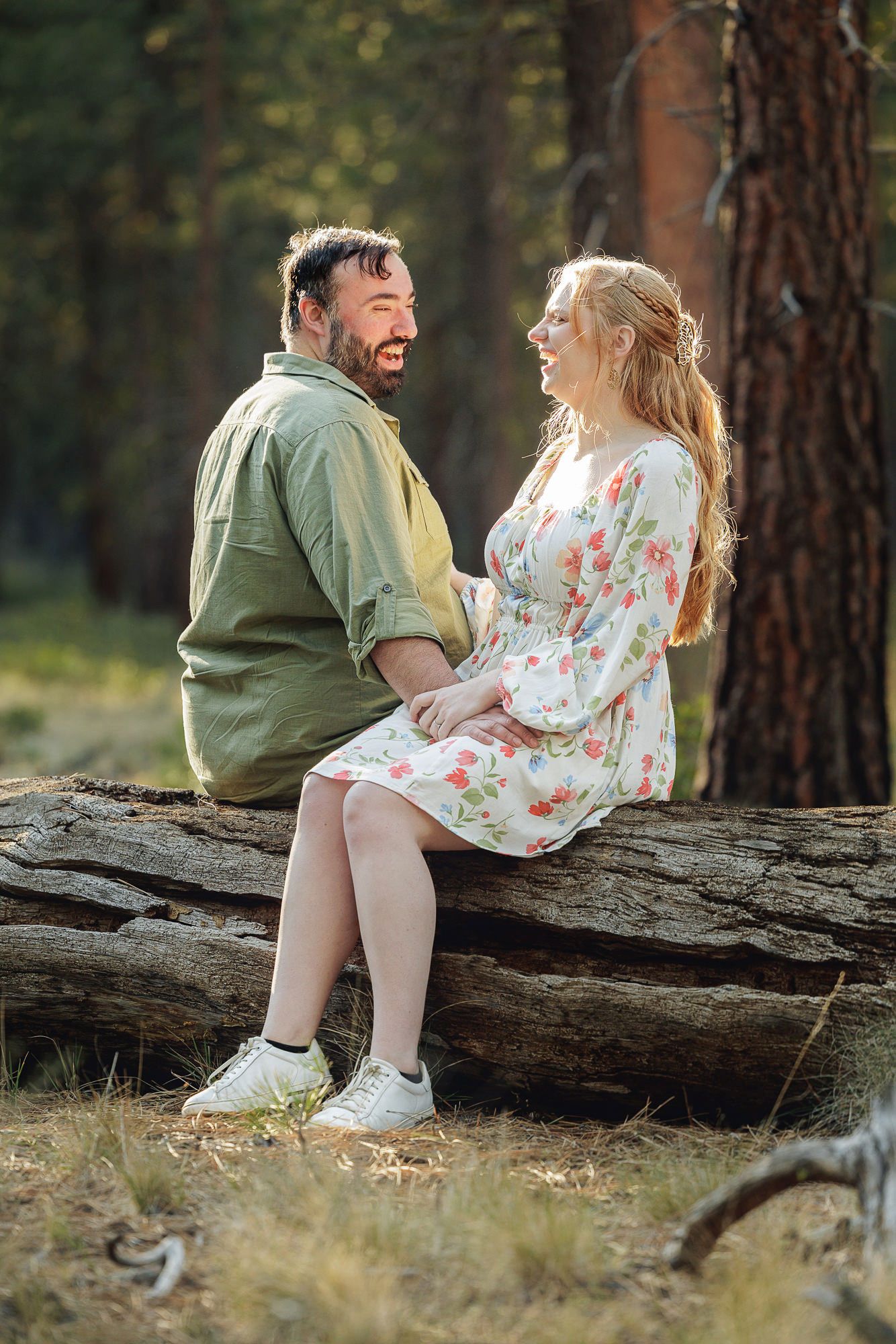 A smiling couple sits together on a fallen log in a sunlit forest, the woman wearing a floral dress and the man in a green shirt.