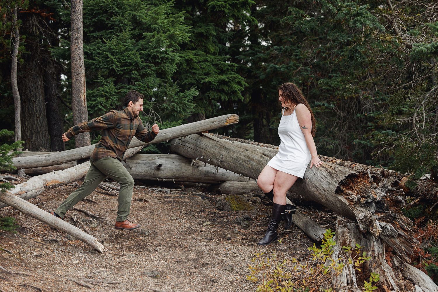 Someone climbs over fallen logs on a forest trail while wearing casual outdoor clothing.