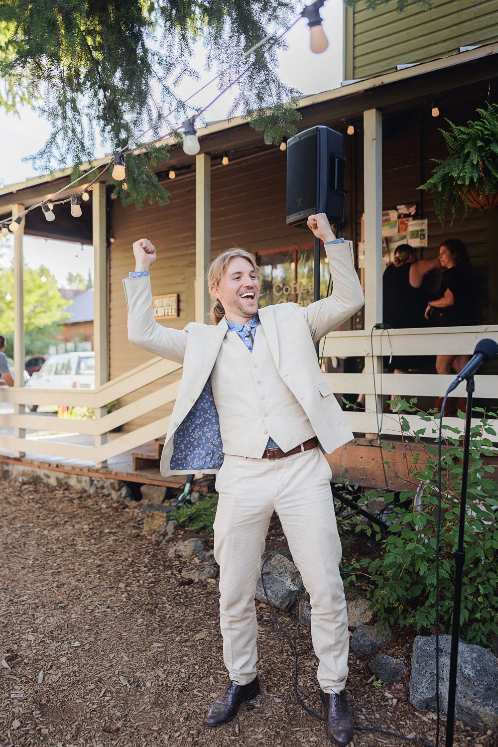 A person in a white suit strikes playful poses while celebrating on a wooden porch with string lights and greenery.