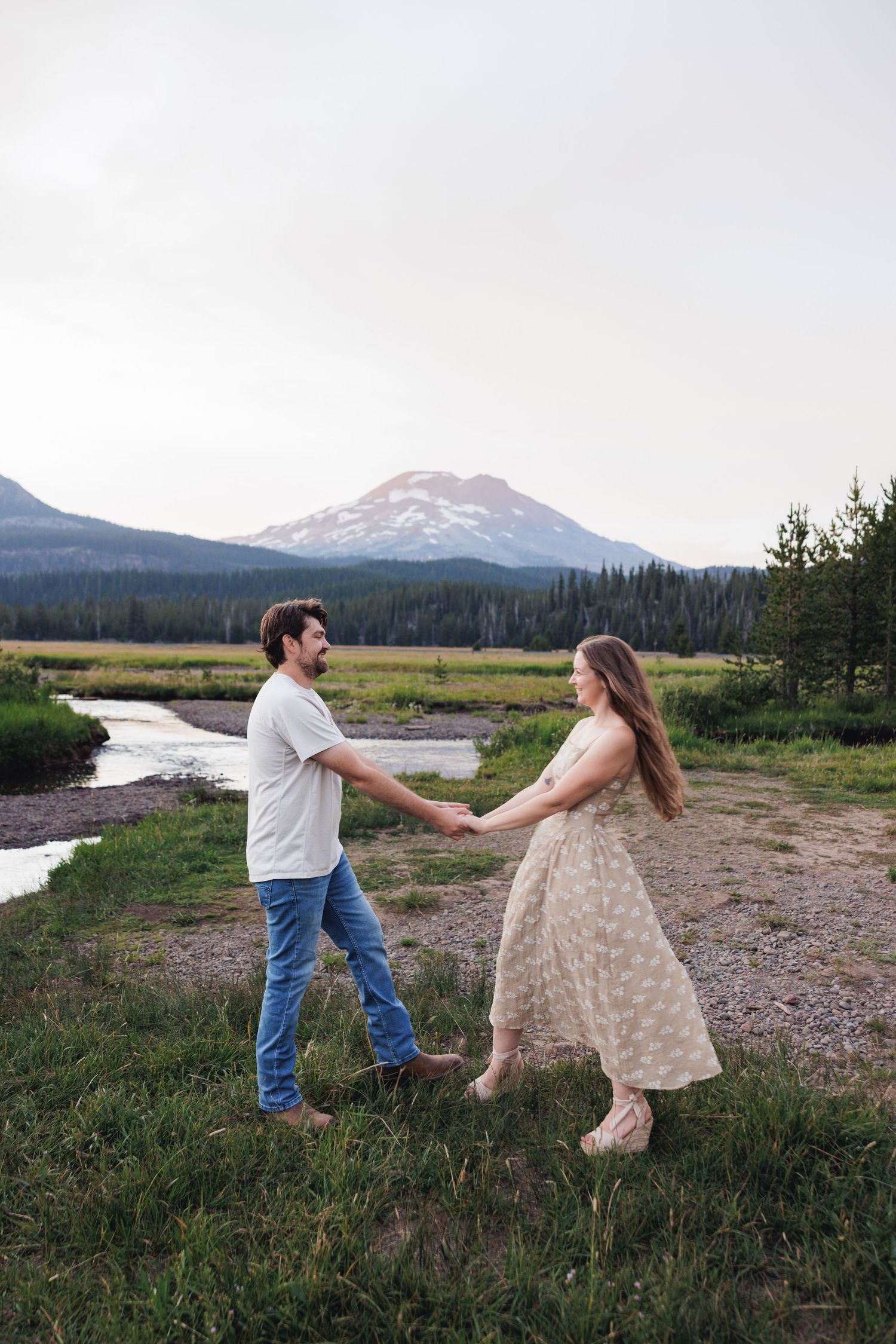 A couple dances together in a meadow with a majestic snow-capped mountain and winding river in the background.