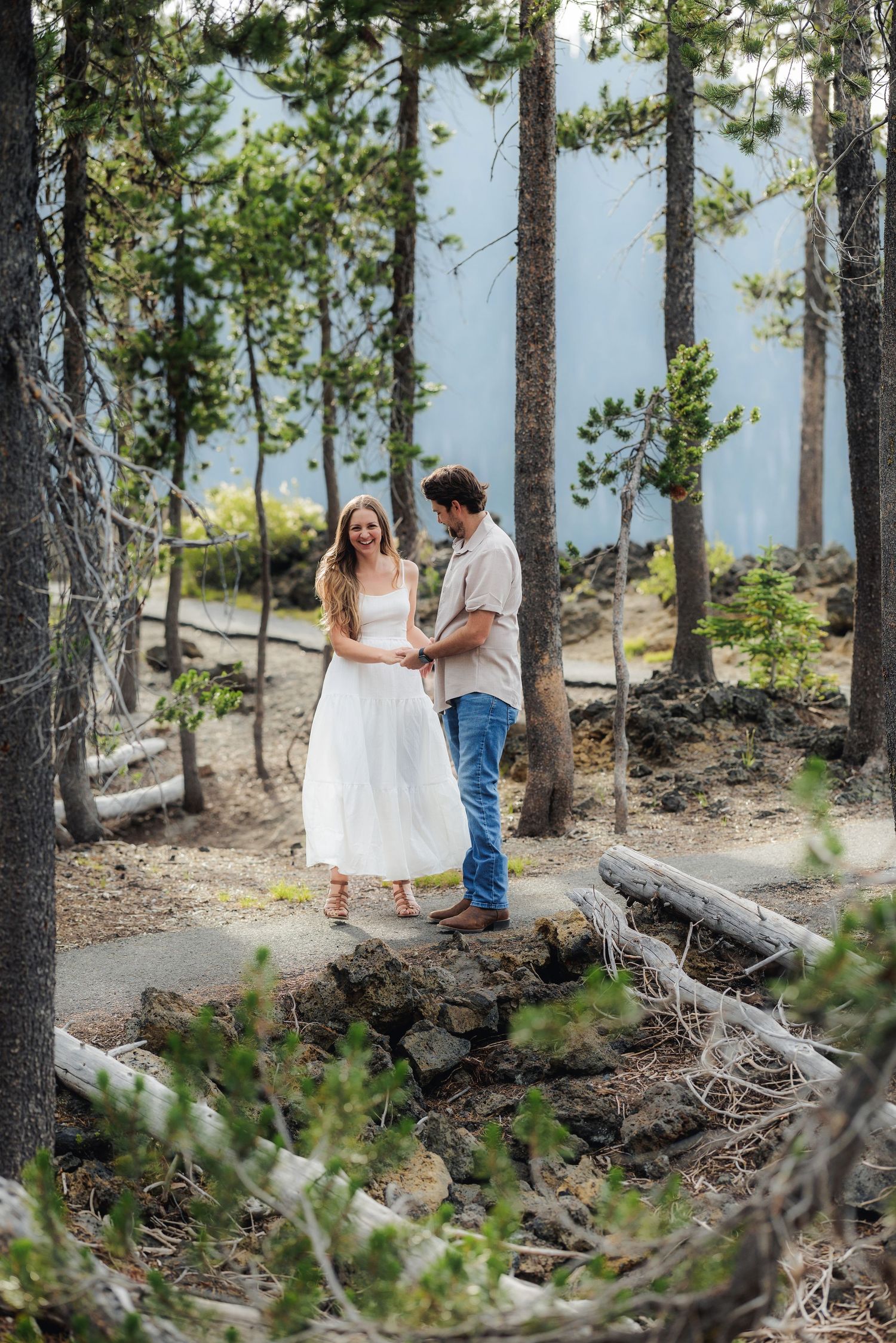 A couple walks together through a scenic forest path among tall pine trees near a mountainous overlook.
