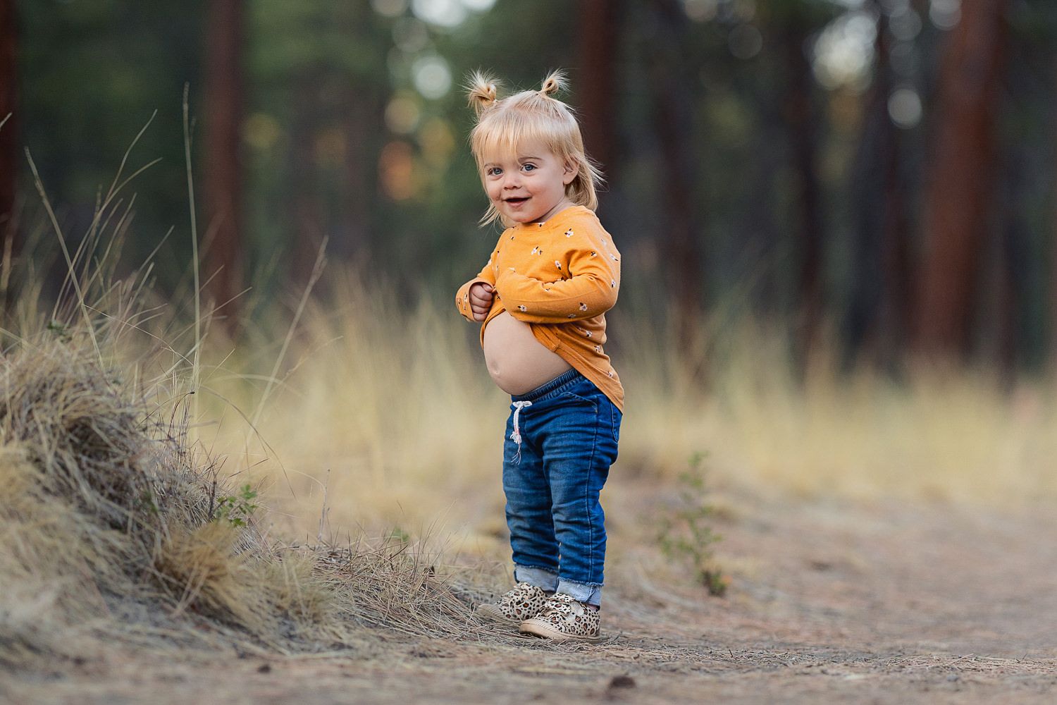 Small child in orange sweater and jeans stands on forest trail during autumn afternoon.