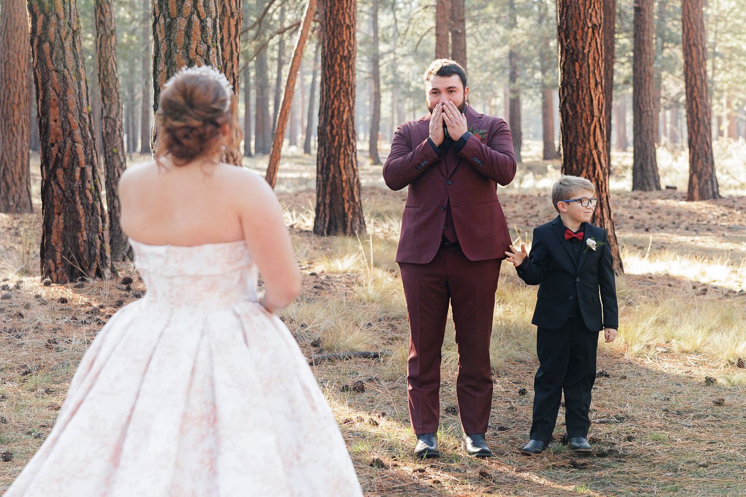 A wedding couple shares a romantic moment among tall pine trees in a sunlit forest setting.