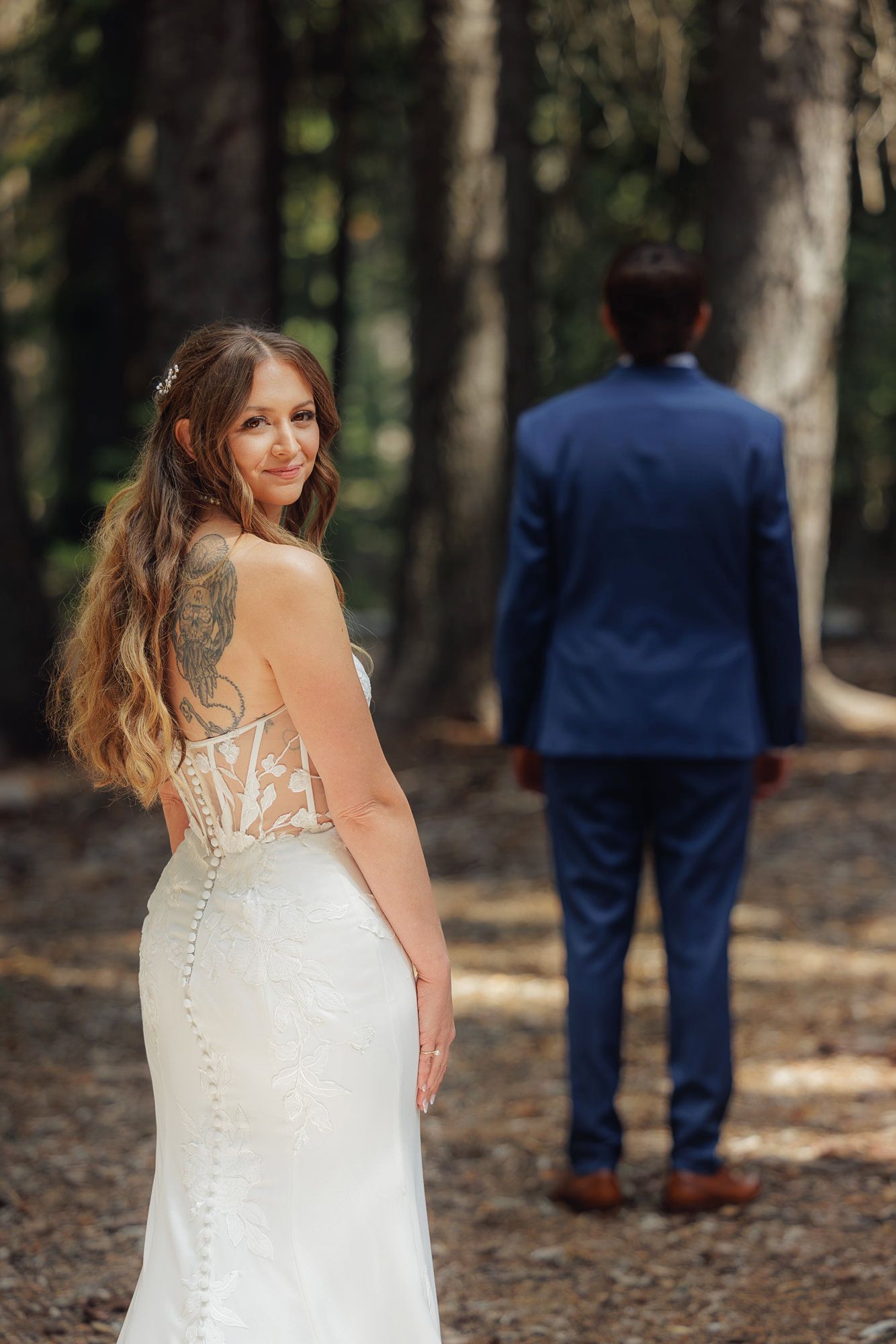 A bride in a white lace dress looks back over her shoulder while standing in a wooded forest setting.