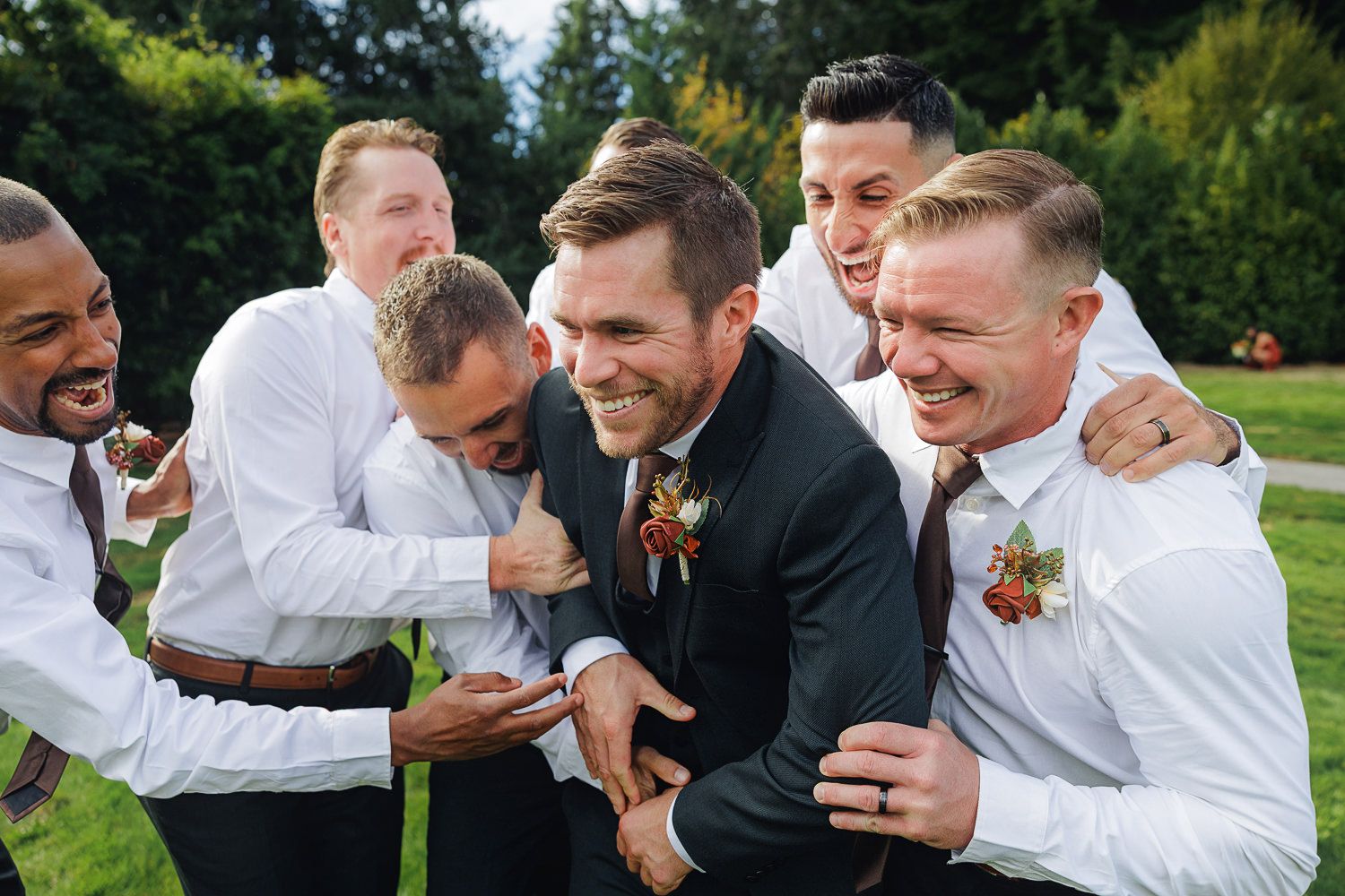 Groomsmen in black suits and white shirts share laughter and hugs during outdoor wedding photo session.
