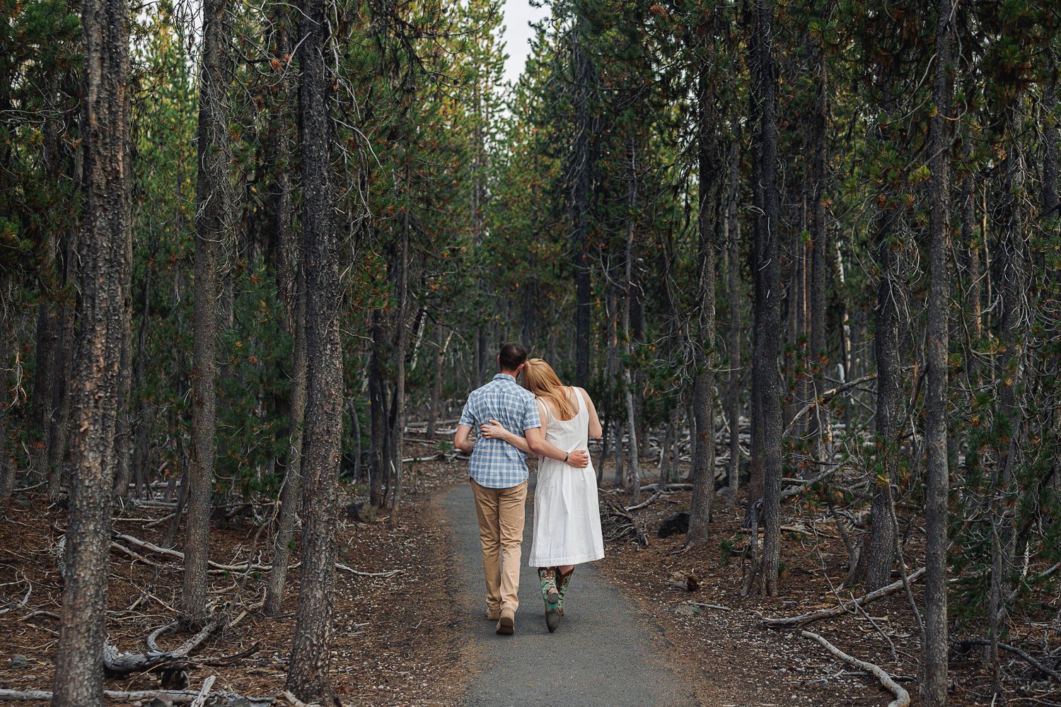 A couple walks holding hands down a rustic dirt path through a dense wooded forest area at dusk.