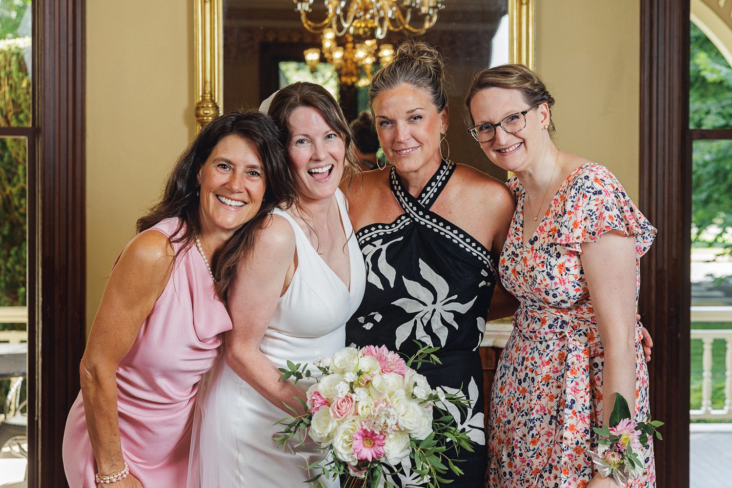 Wedding party wearing pink and floral dresses laugh together in an elegant indoor setting with chandelier and bouquets.