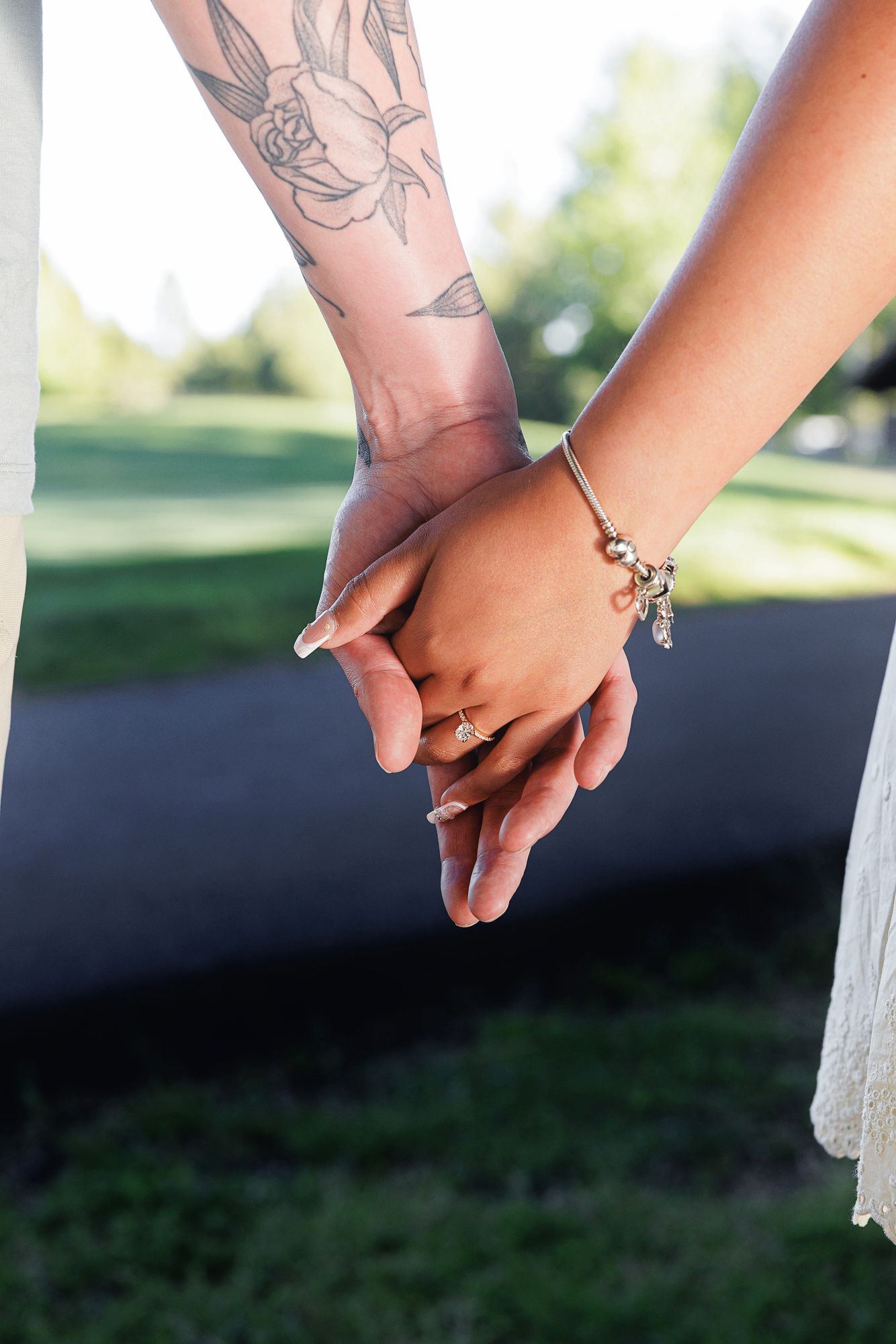 Close-up of two hands holding, one with floral tattoos and one wearing a charm bracelet and diamond ring, outdoors.