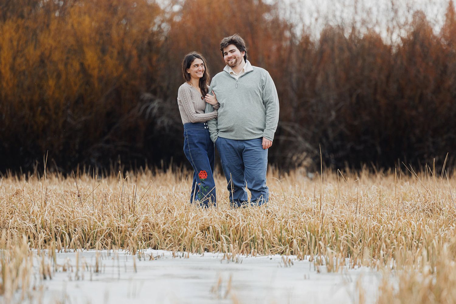 Couple walking together through a golden autumn field during sunset while holding hands and looking at each other.