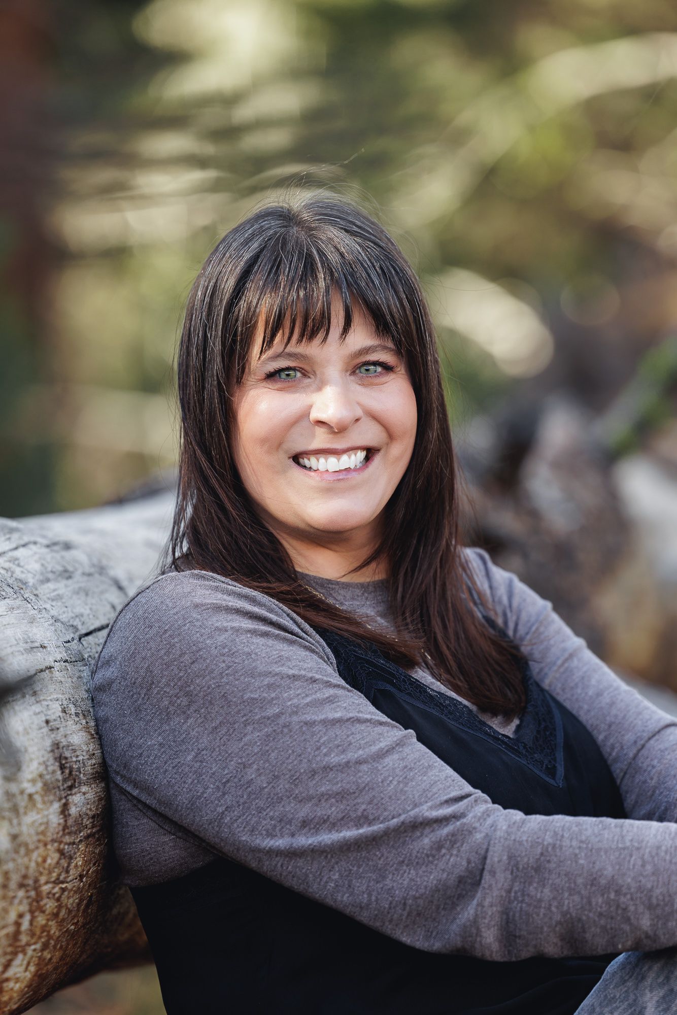 Smiling woman with dark hair and bangs posing outdoors against a blurred natural background.
