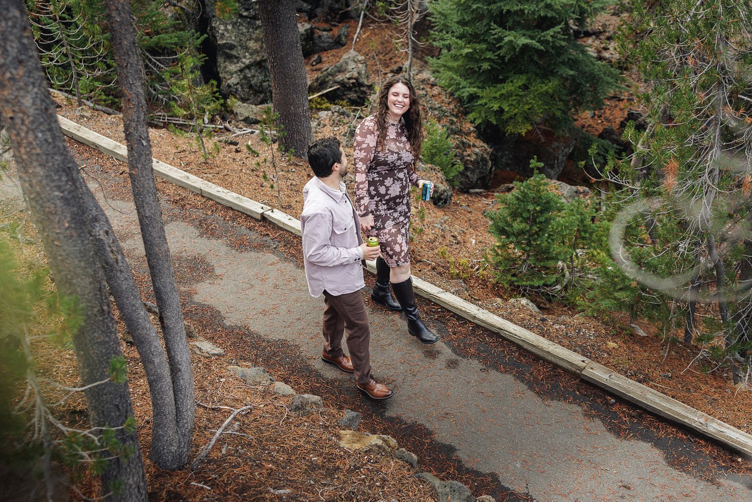 A person walks along a winding forest path surrounded by tall trees and scattered autumn leaves on the ground.