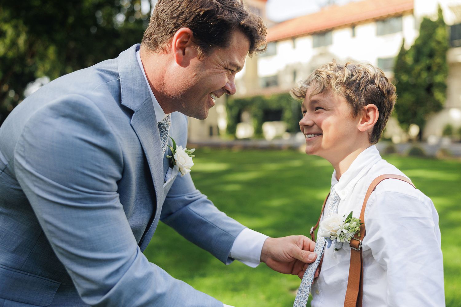 A person in a blue suit helps a child adjust their outfit while outdoors in a garden setting.