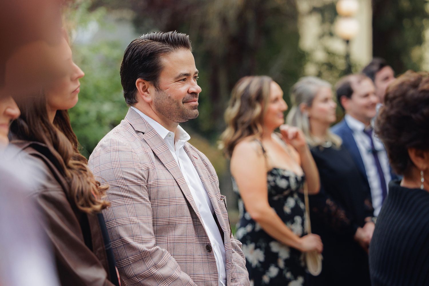 Wedding guests smile and laugh during an outdoor ceremony in formal attire.