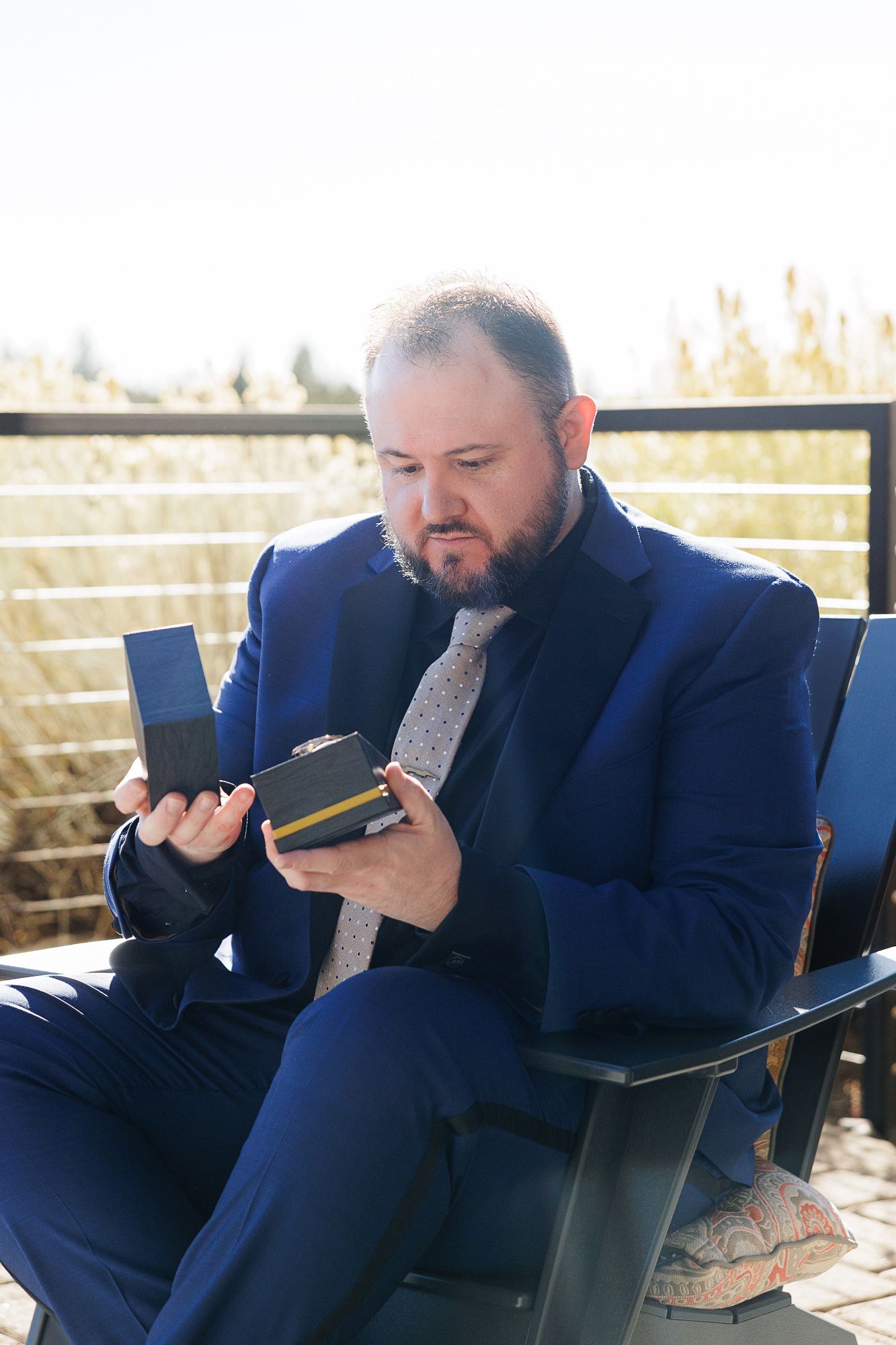 A person in a navy suit sits on a patio and looks at their phone while holding a beverage.