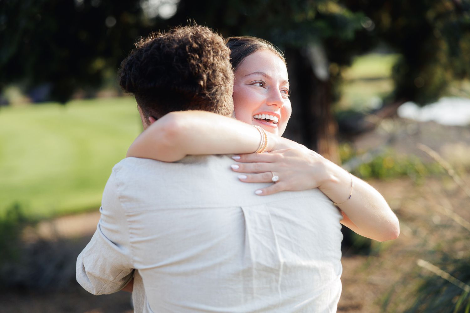 A smiling bride hugs her groom outdoors in soft natural light, wearing a white dress with a ring visible on her hand.