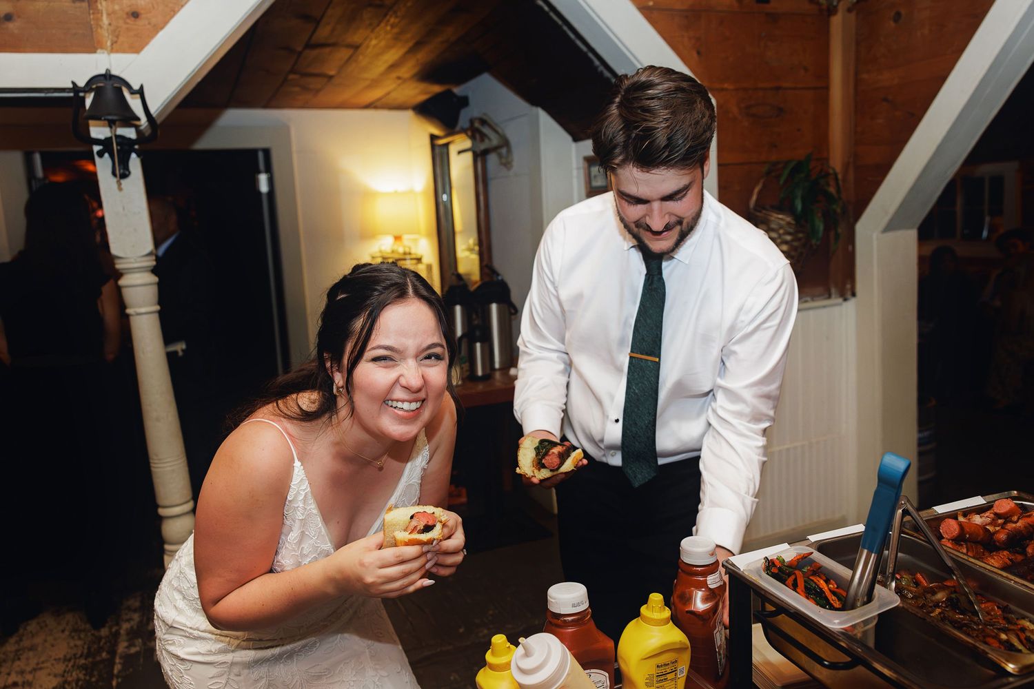 Bride and groom laughing together at a food station during their wedding reception in a rustic barn venue.
