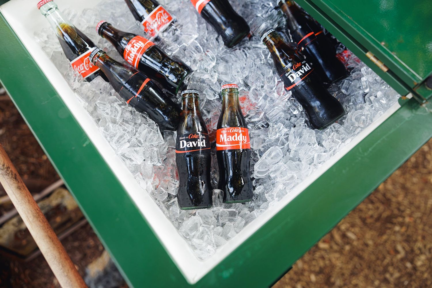 Personalized Coca-Cola glass bottles chilling on ice in a green cooler at an outdoor event.