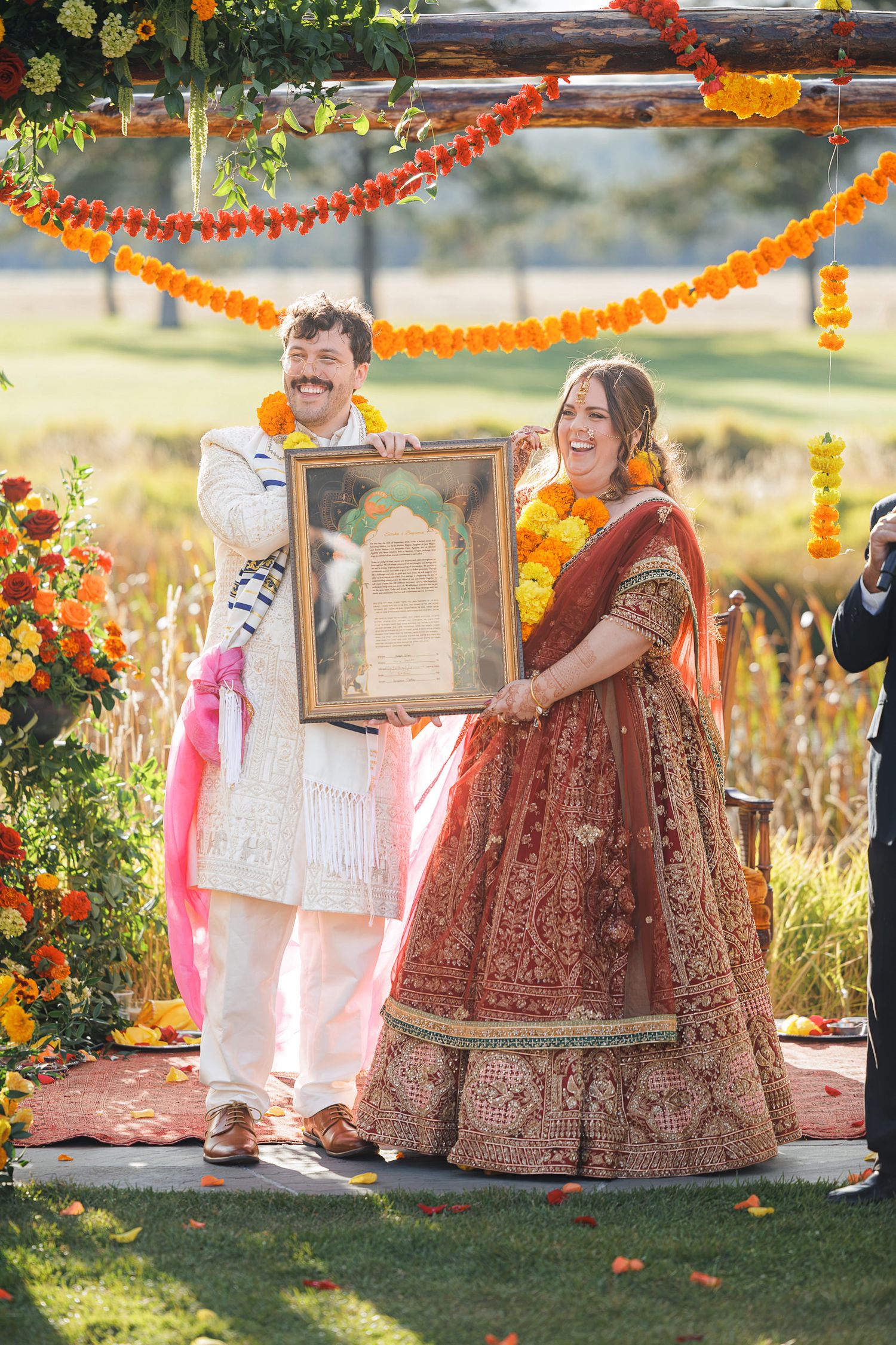 Couple in traditional Indian wedding attire holding framed vows under marigold garland mandap outdoors.