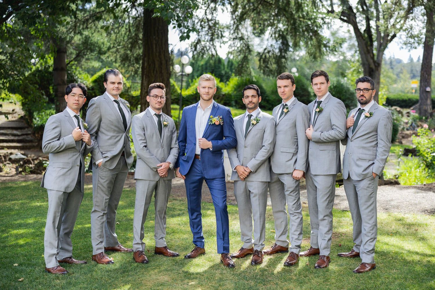 A group in formal attire poses outdoors, with one wearing a blue suit among others in light gray suits in a garden setting.