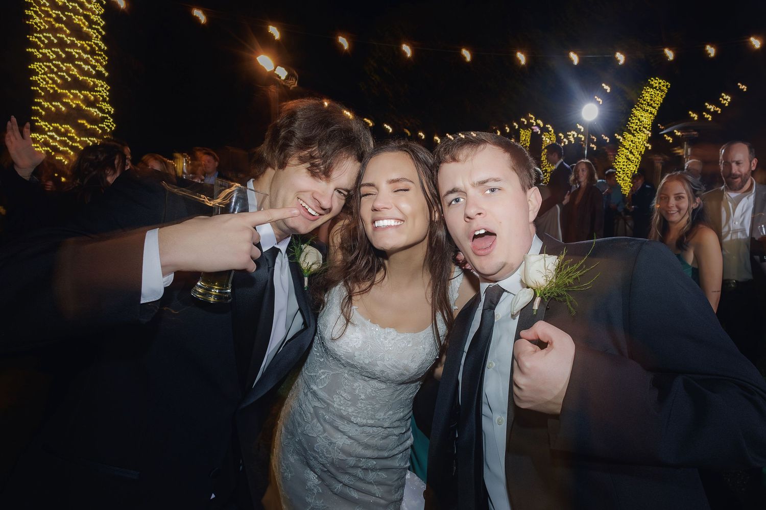 Wedding party celebrates with energetic dancing at a festive outdoor nighttime reception with string lights overhead.